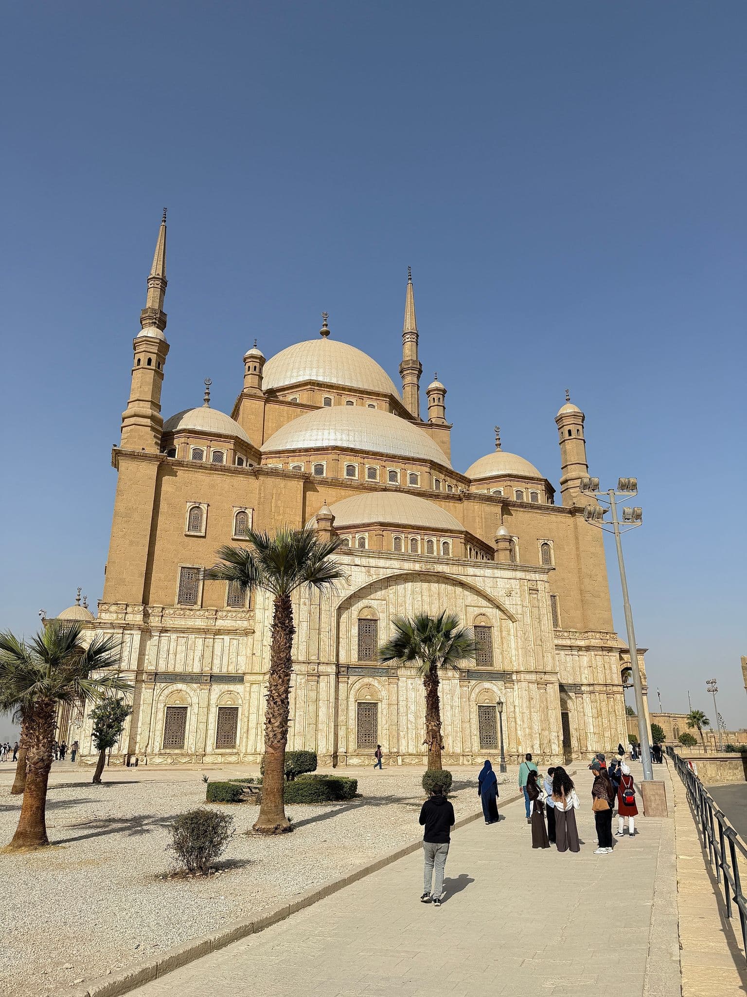 Mosque of Muhammad Ali at the Citadel in Cairo, Egypt, with visitors walking on the forecourt under a clear sky.
