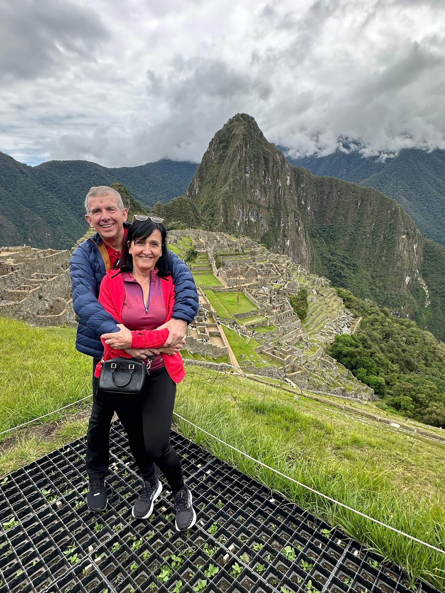 Machu Picchu terraces with Huayna Picchu behind and two travelers hugging on a grassy hillside, Cusco Region, Peru.