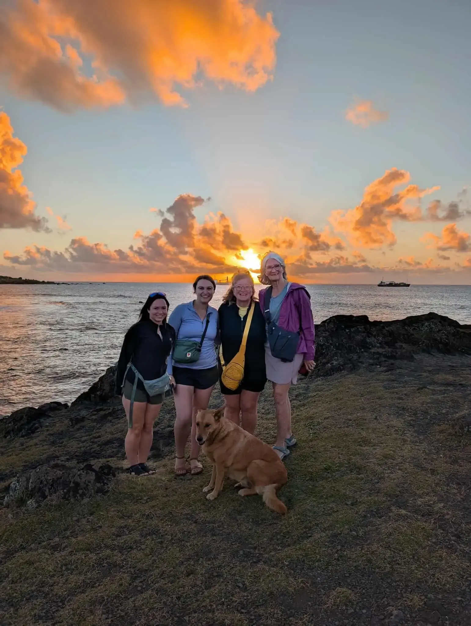 Easter Island coastline at sunset with four travelers standing on rocky shore and a tan dog sitting in front, Rapa Nui, Chile.