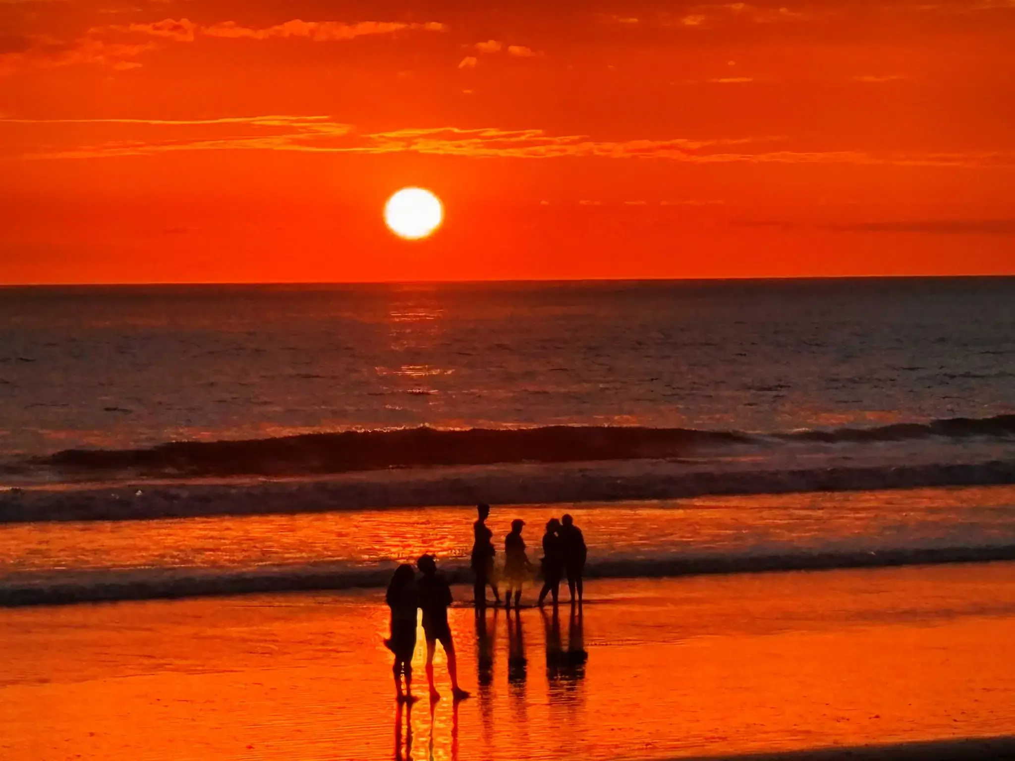 Sunset over a Costa Rica beach with the sun near the horizon and silhouetted people standing in shallow water.