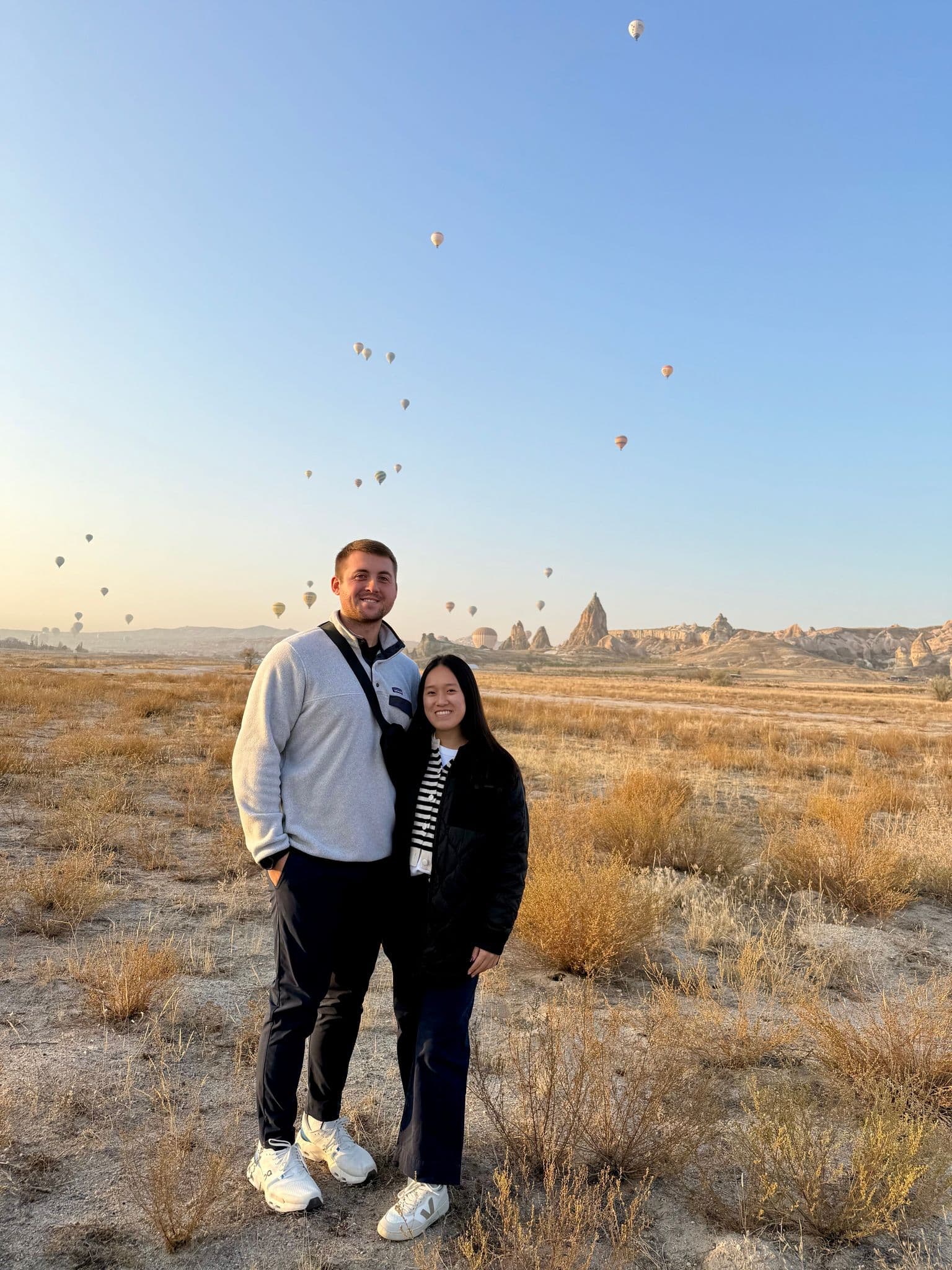 Hot air balloons floating over Cappadocia rock formations with a couple posing in the foreground, Cappadocia, Turkey.
