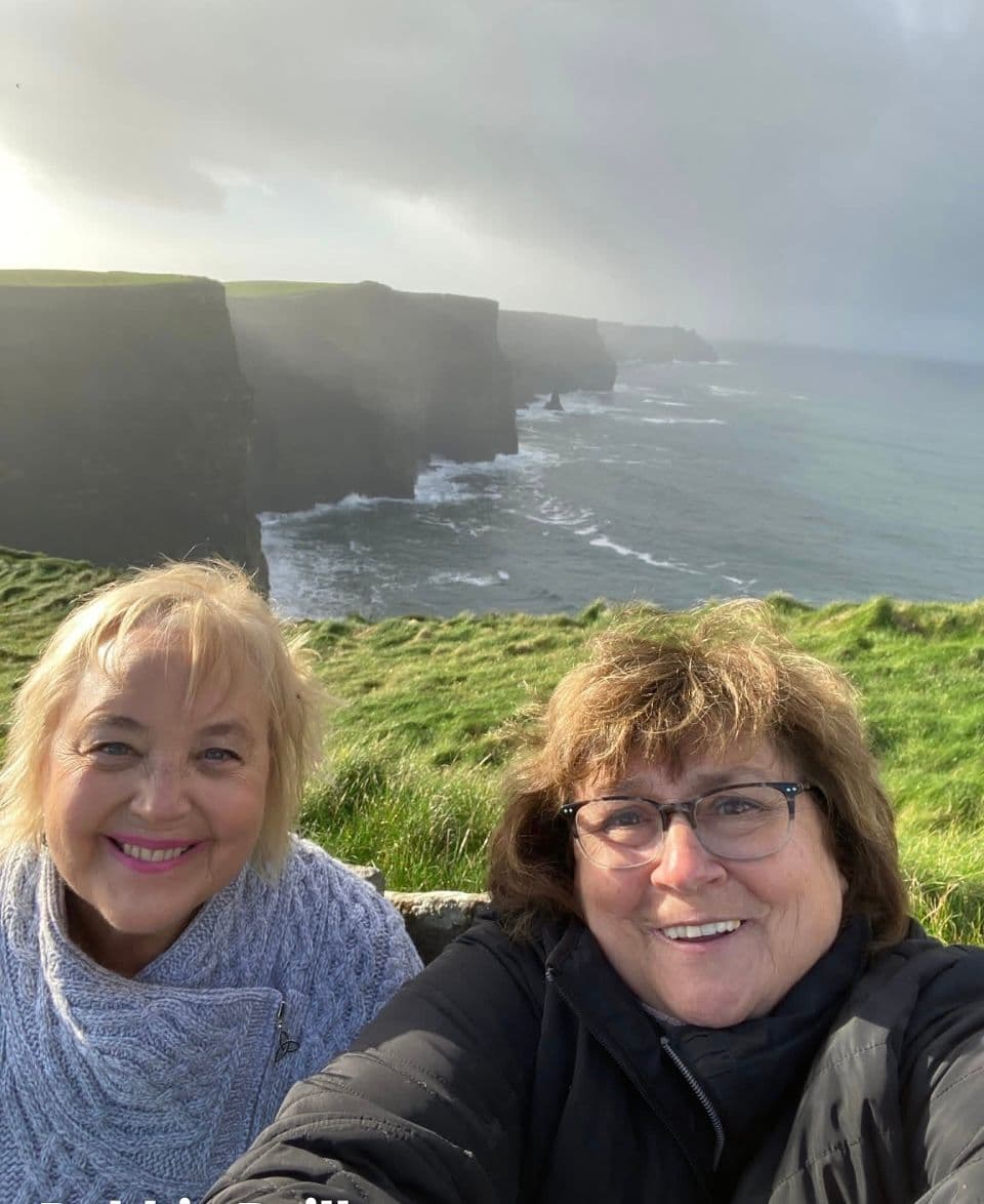 Cliffs of Moher and Atlantic coastline with two women taking a selfie on the grassy cliff edge, County Clare, Ireland.