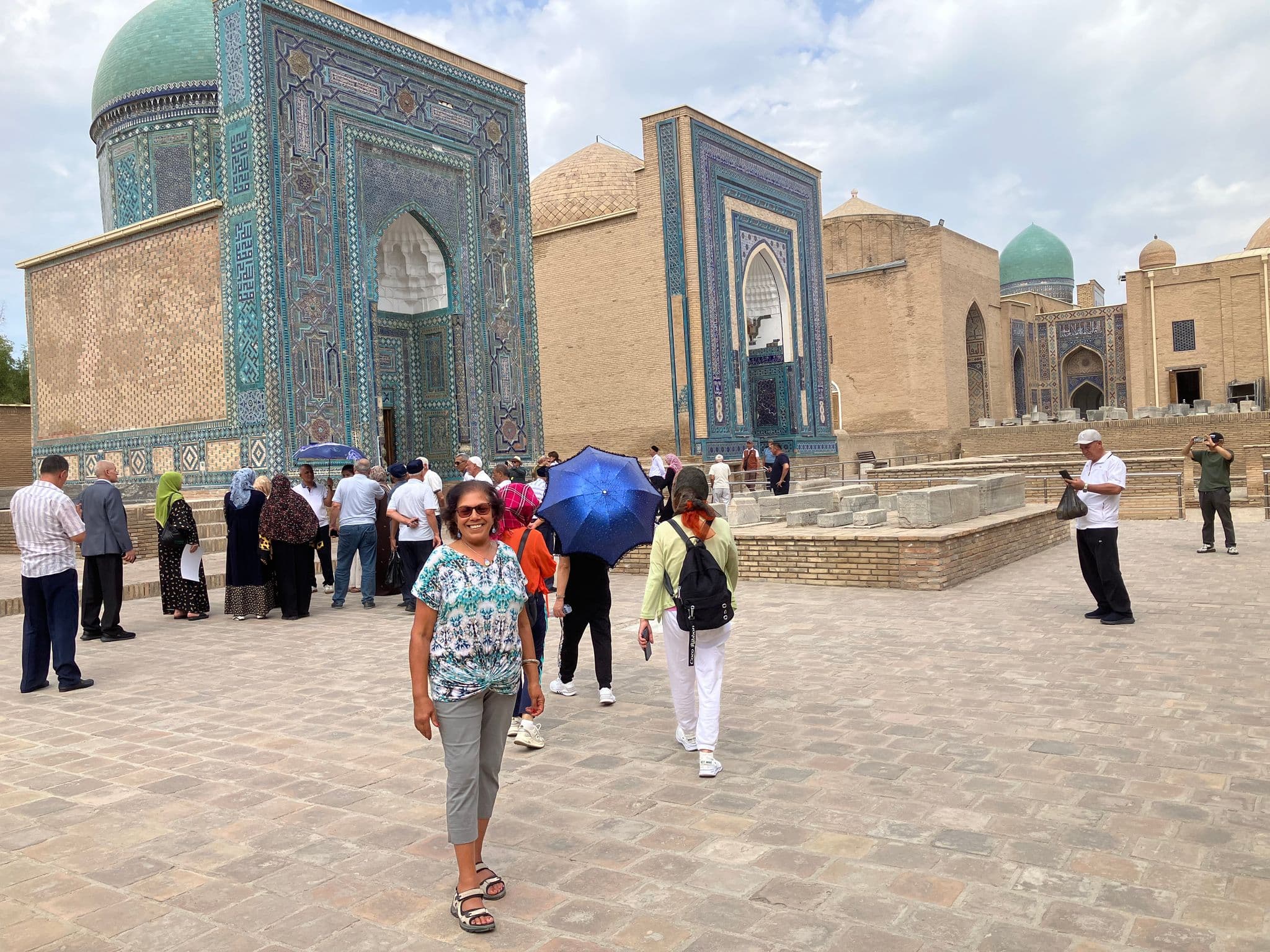 Shah-i-Zinda mausoleums in Samarkand, Uzbekistan, with a woman standing and smiling in the courtyard among other tourists.