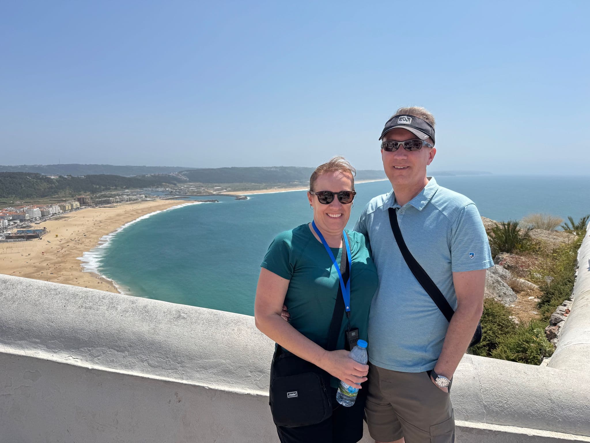 Praia da Nazaré crescent beach from a cliff viewpoint with two travelers posing in the foreground, Nazaré, Portugal.
