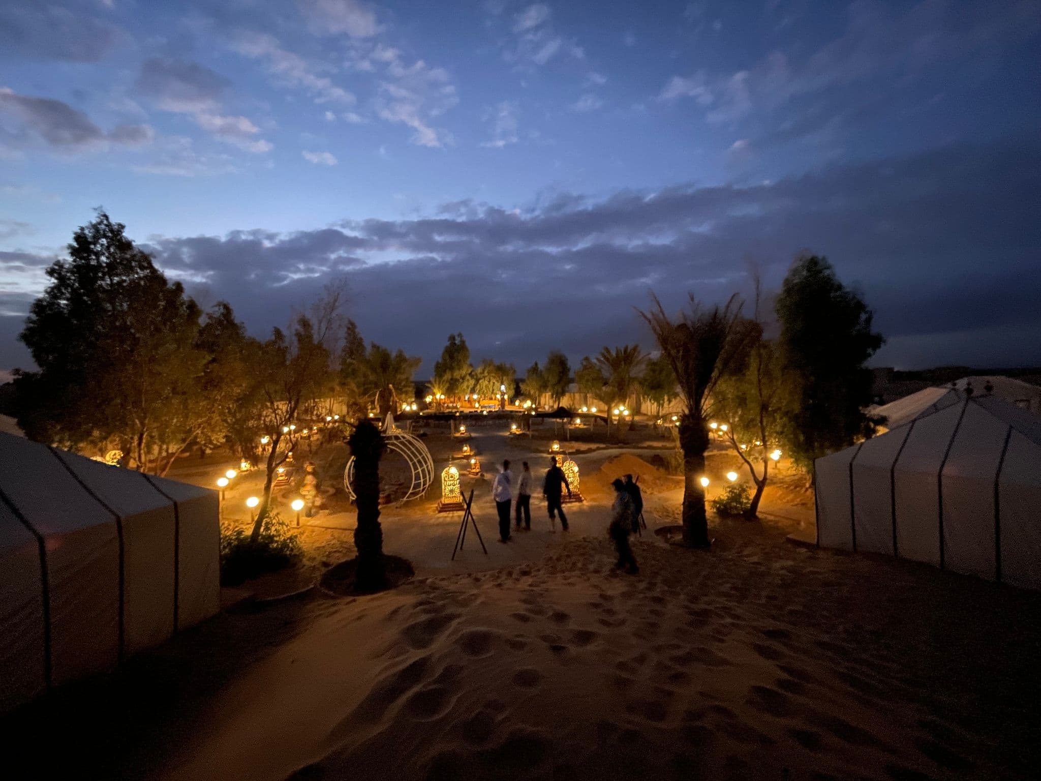 Lit desert camp with tents, lanterns, and people walking across sand at dusk, Sahara Desert.