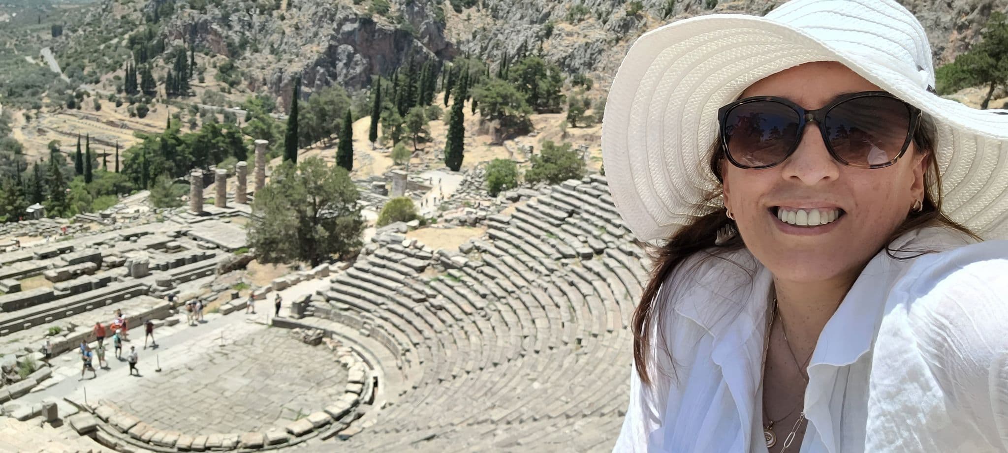 Temple of Apollo ruins and stone theater at Delphi, Greece, with a woman in a white sunhat taking a selfie.