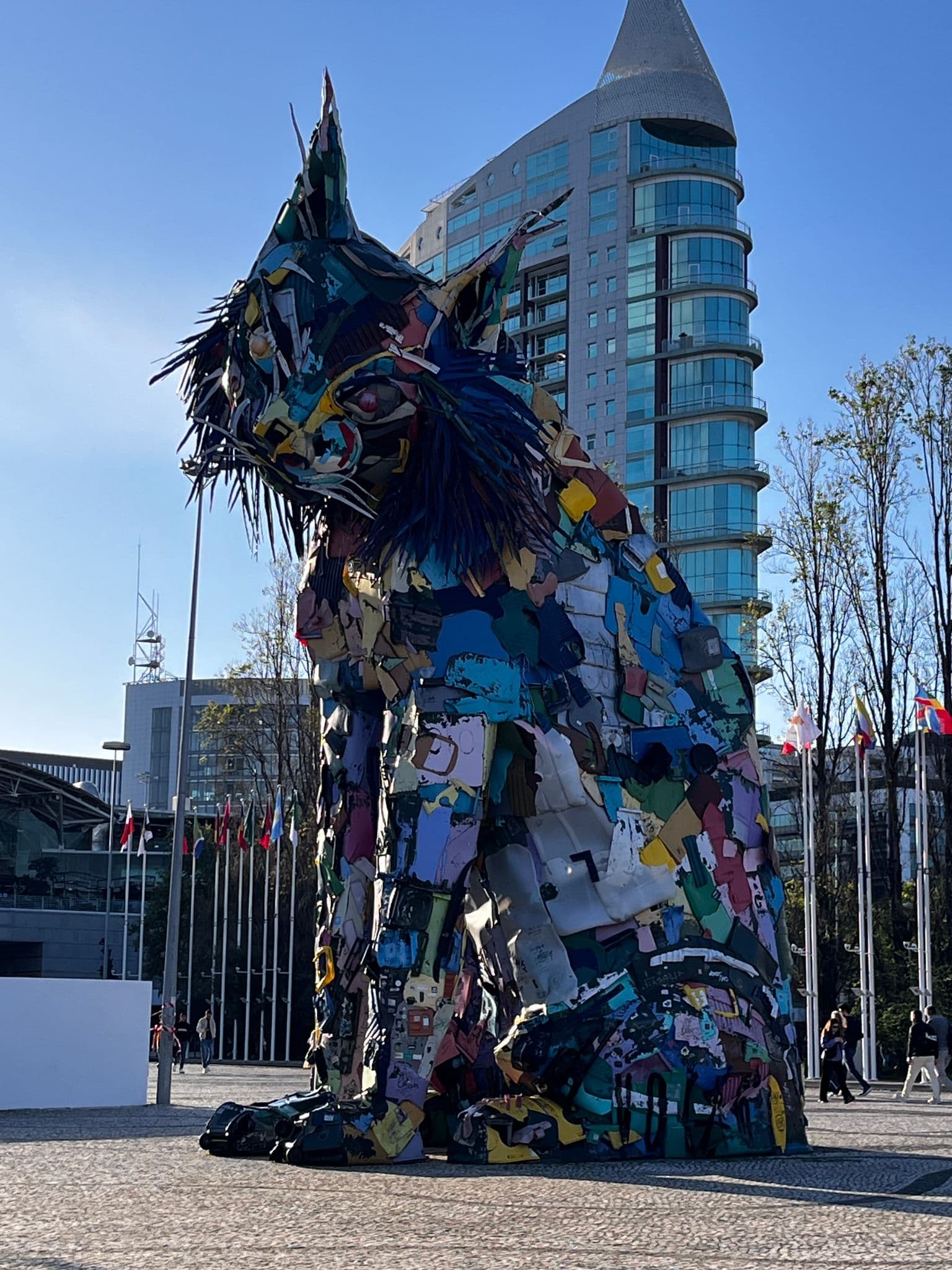 Large colorful lynx sculpture made from recycled plastic in a Lisbon plaza with modern glass buildings and flagpoles in Portugal.