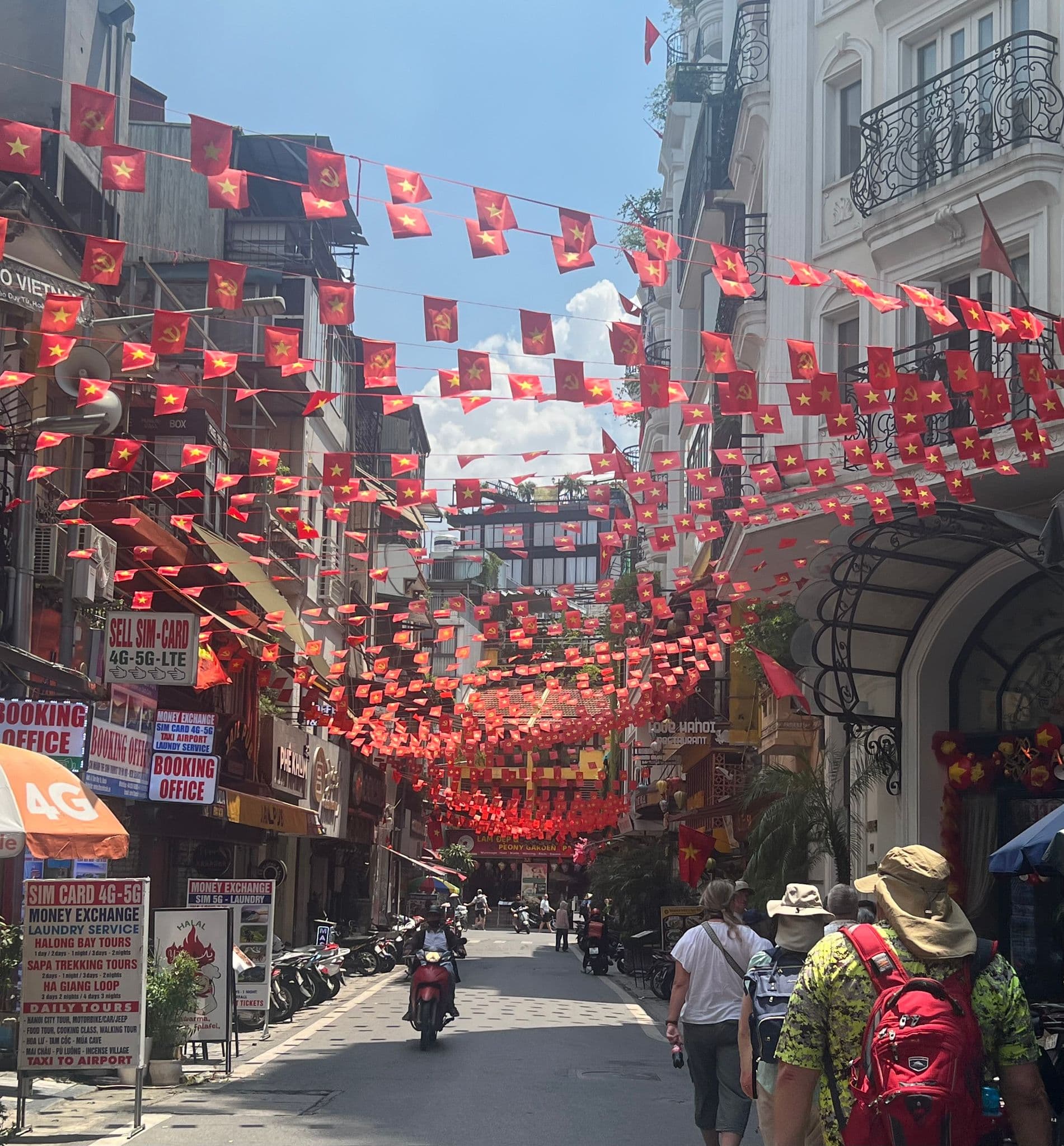 Rows of Vietnamese flags hung across a narrow street in the Hanoi Old Quarter, Hanoi, Vietnam, with tourists walking and a motorbike passing through.