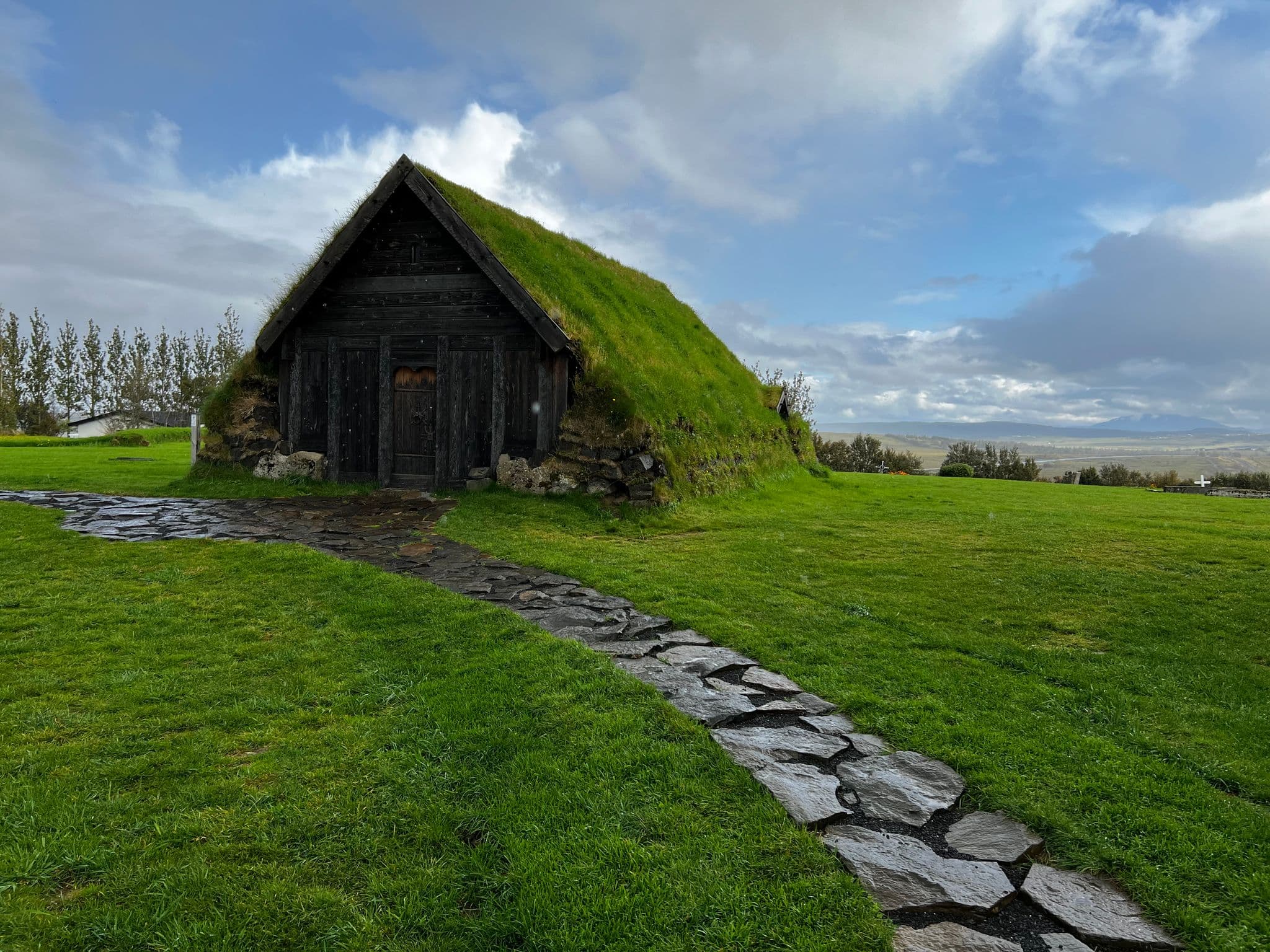 Turf-roofed chapel with a stone path leading to its door on a grassy hill in rural Iceland.