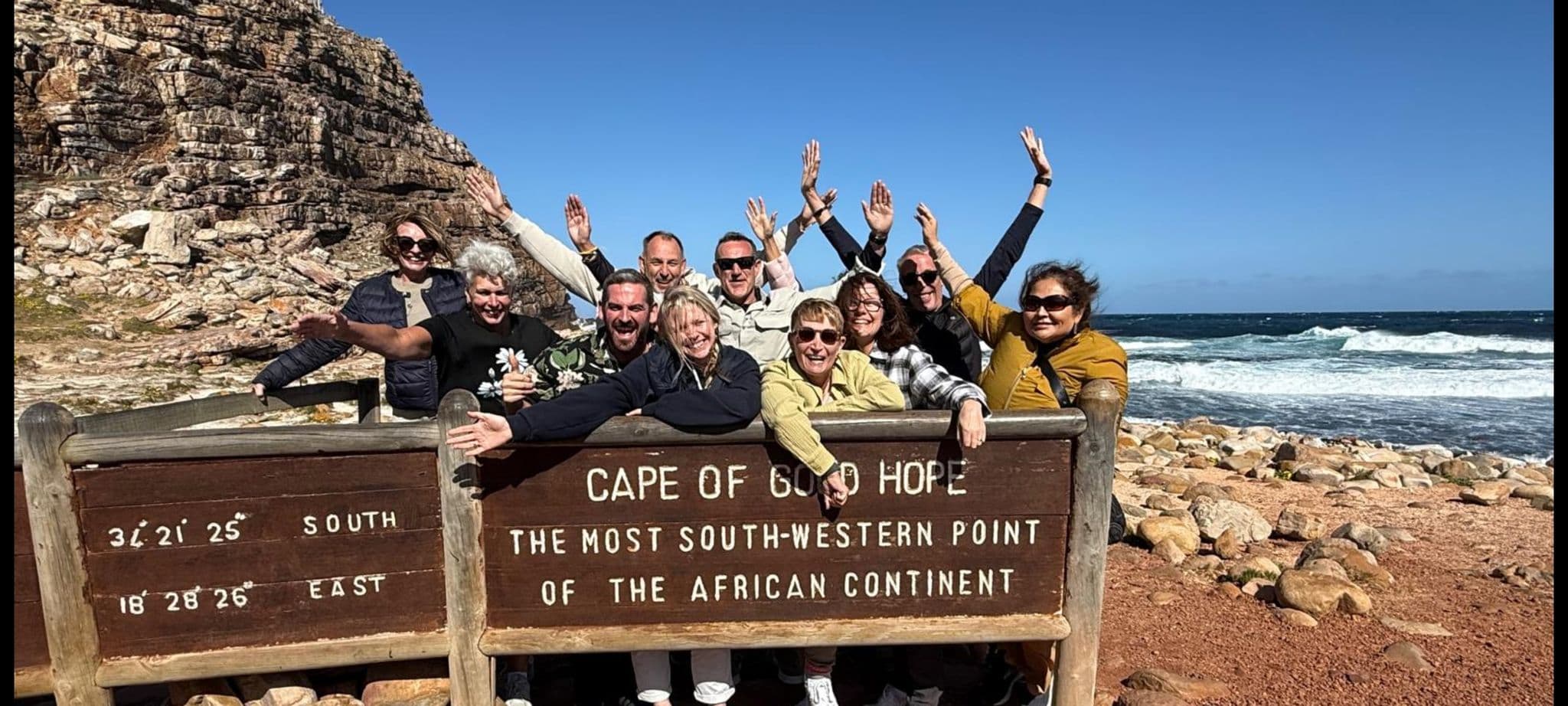 Cape of Good Hope sign with a group of travelers leaning on it and cheering by the rocky shore in South Africa.