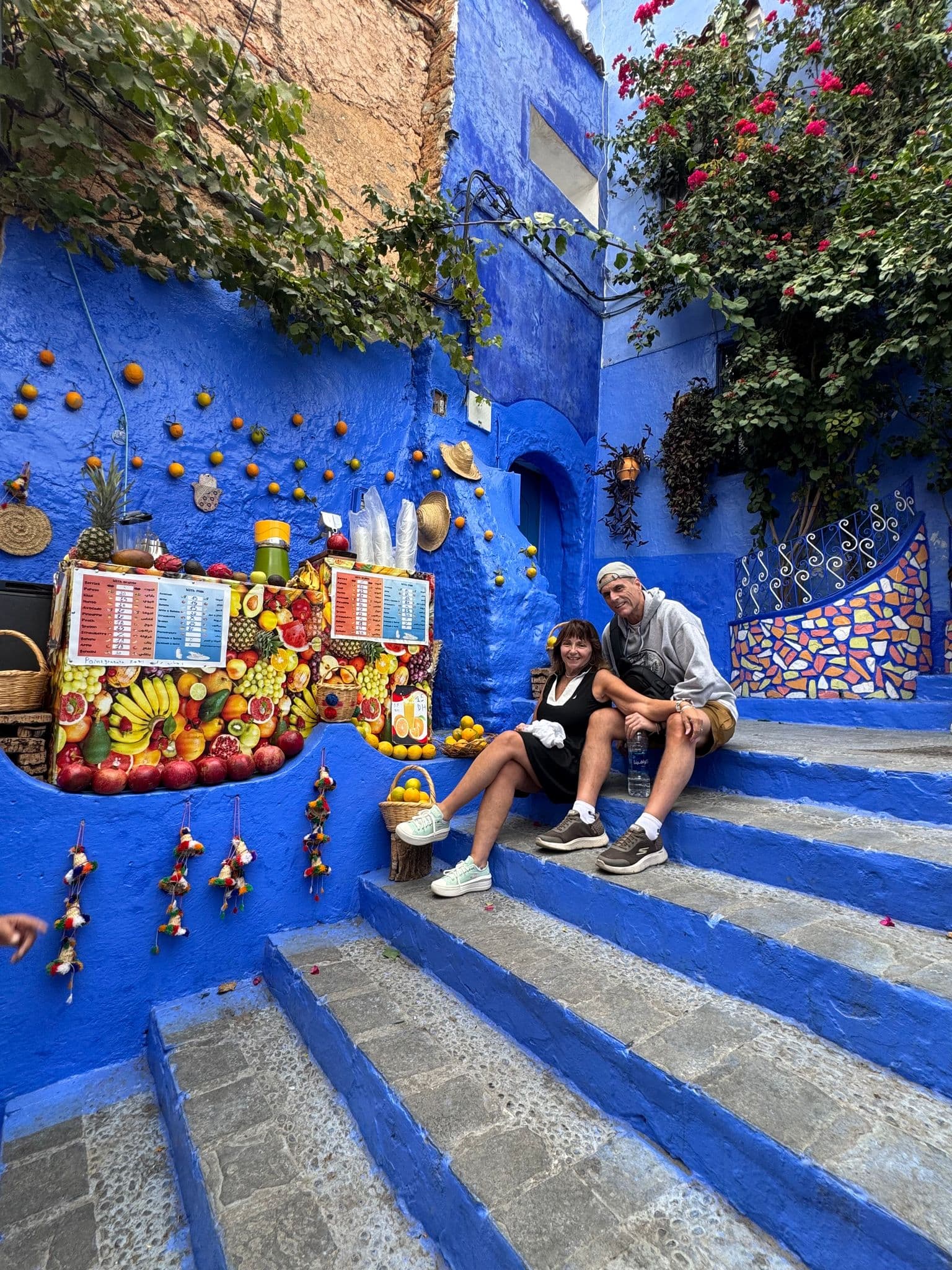 Blue-painted steps and walls in Chefchaouen, Morocco, with two travelers sitting on the stairs beside a colorful fruit and juice stall.