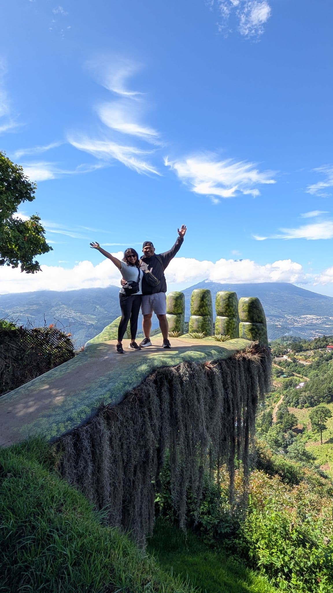 Hobbitenango hand-shaped viewpoint with two people standing and waving on a moss-covered platform overlooking mountains in Guatemala.