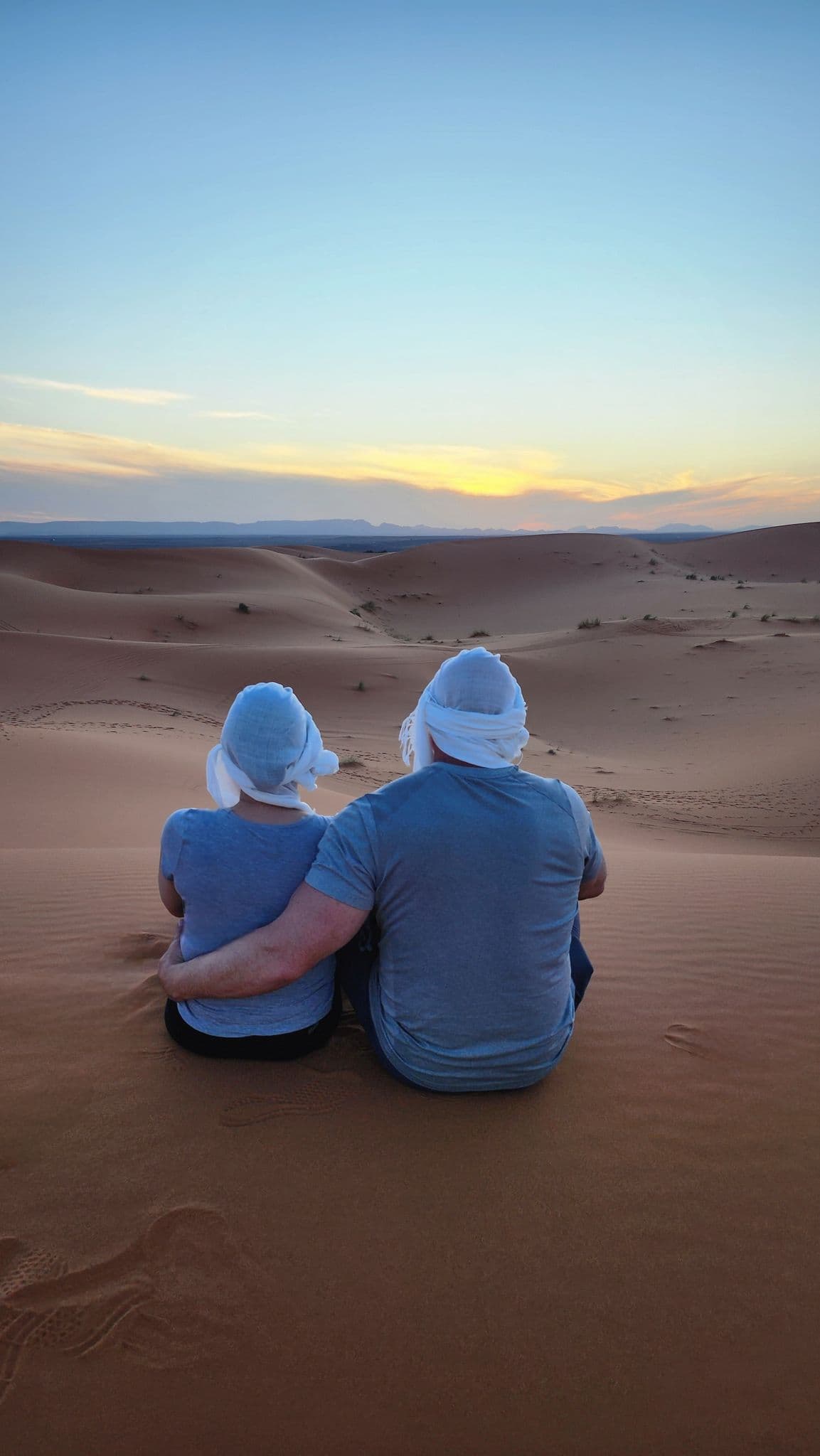 Erg Chebbi sand dunes at sunset with two travelers sitting side-by-side watching the horizon, Merzouga, Morocco.