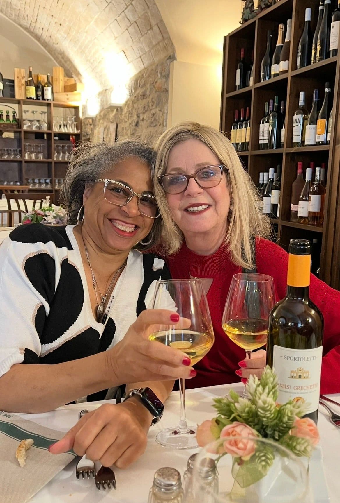 Two women clinking glasses of white wine at a table in a stone-arched restaurant in Italy.