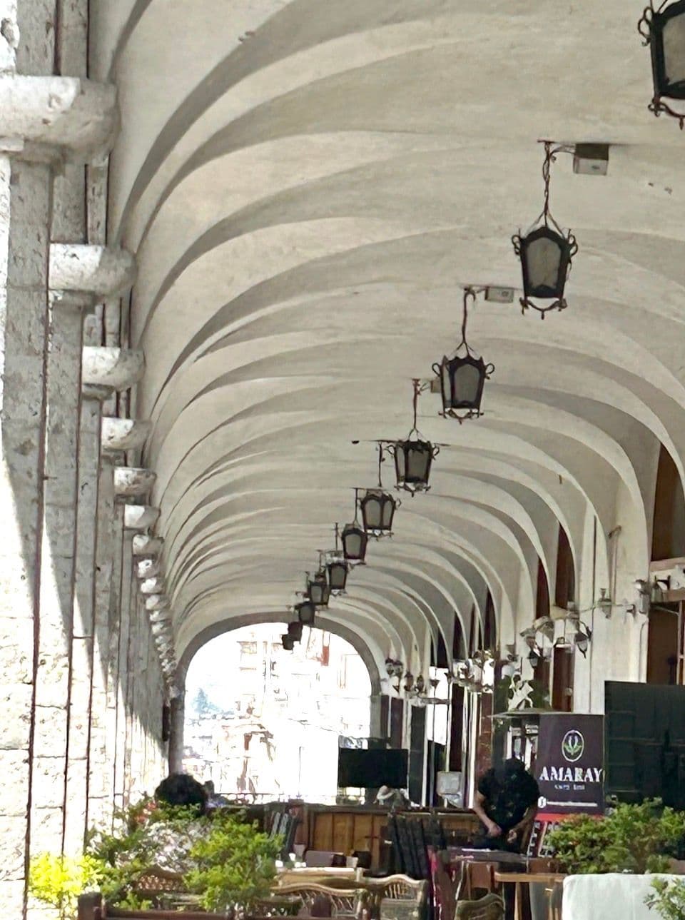 Covered stone arcade of the Plaza de Armas in Cusco, Peru with hanging lanterns and outdoor dining tables.