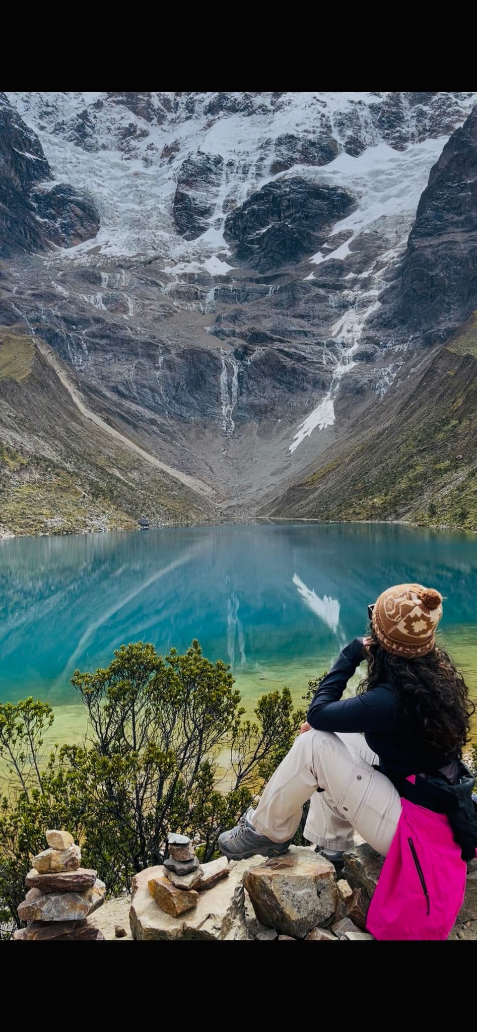 Laguna Humantay with a turquoise glacial lake and steep snow-capped peaks, a hiker sits on rocks looking across the water, Cusco region, Peru.
