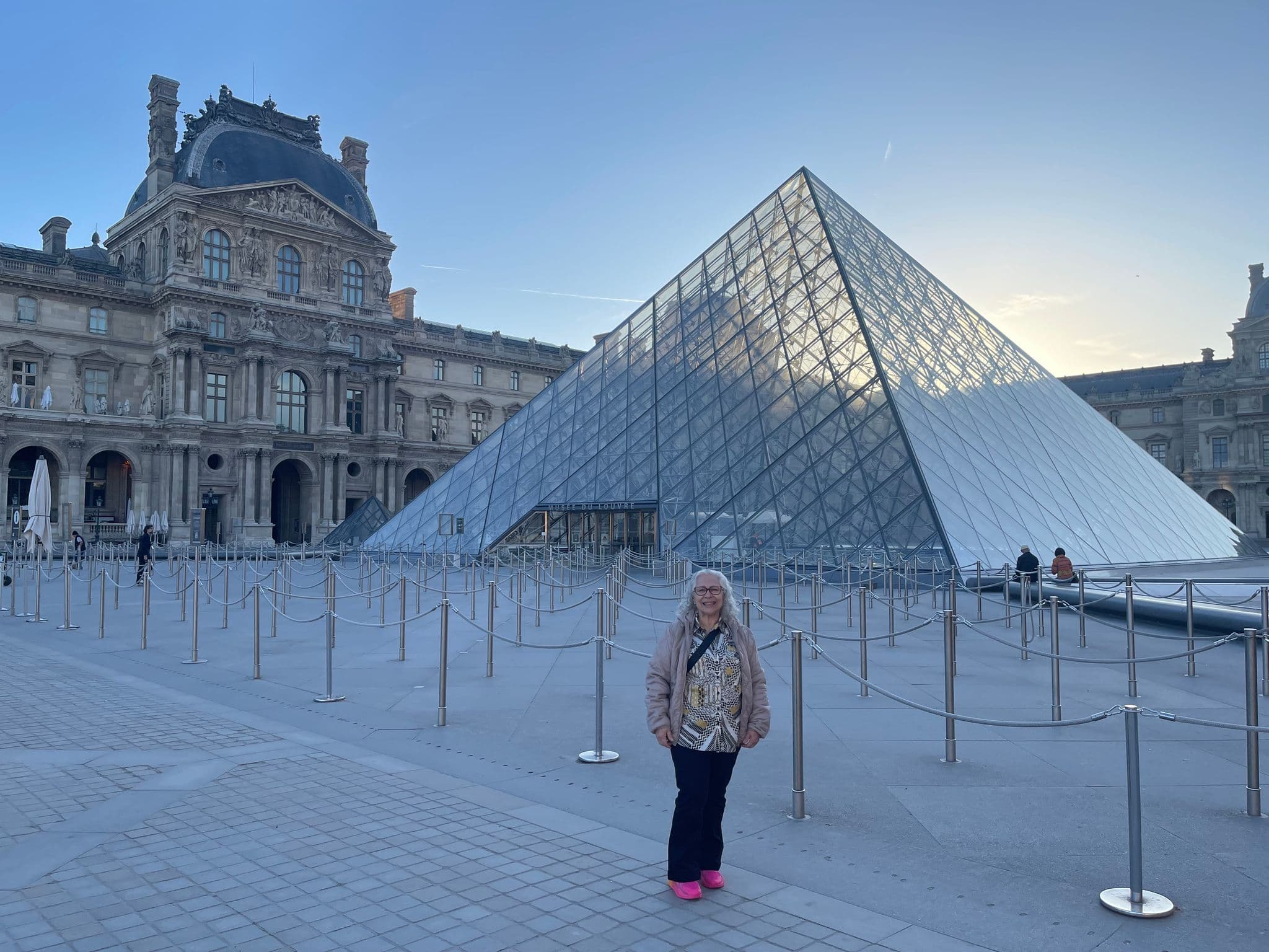 Louvre Pyramid at Musée du Louvre, Paris, France with a traveler standing near queue ropes in the foreground.