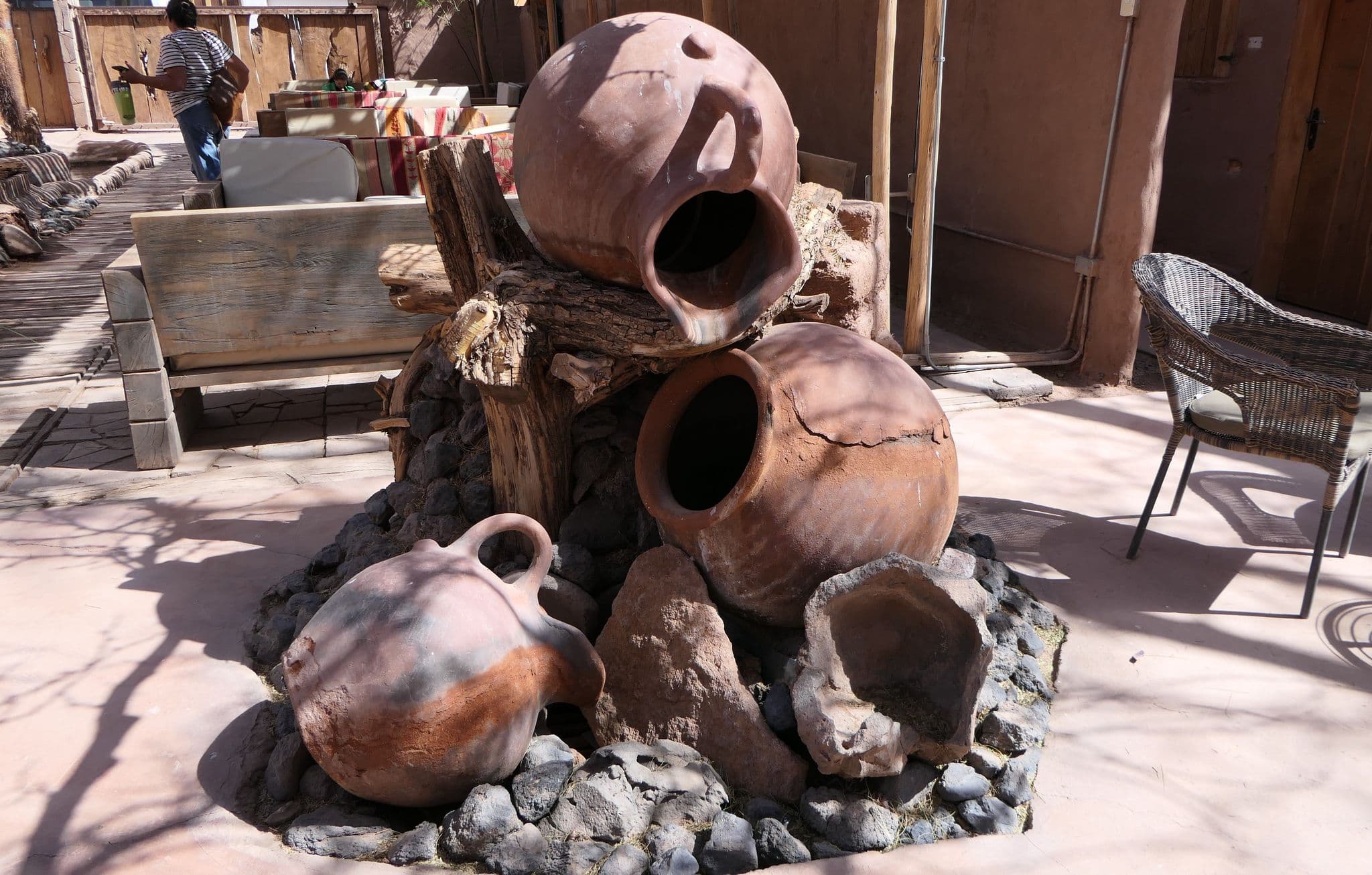 Stacked terracotta jars and broken pots on rocks and driftwood in a hotel courtyard seating area with benches and a wicker chair.