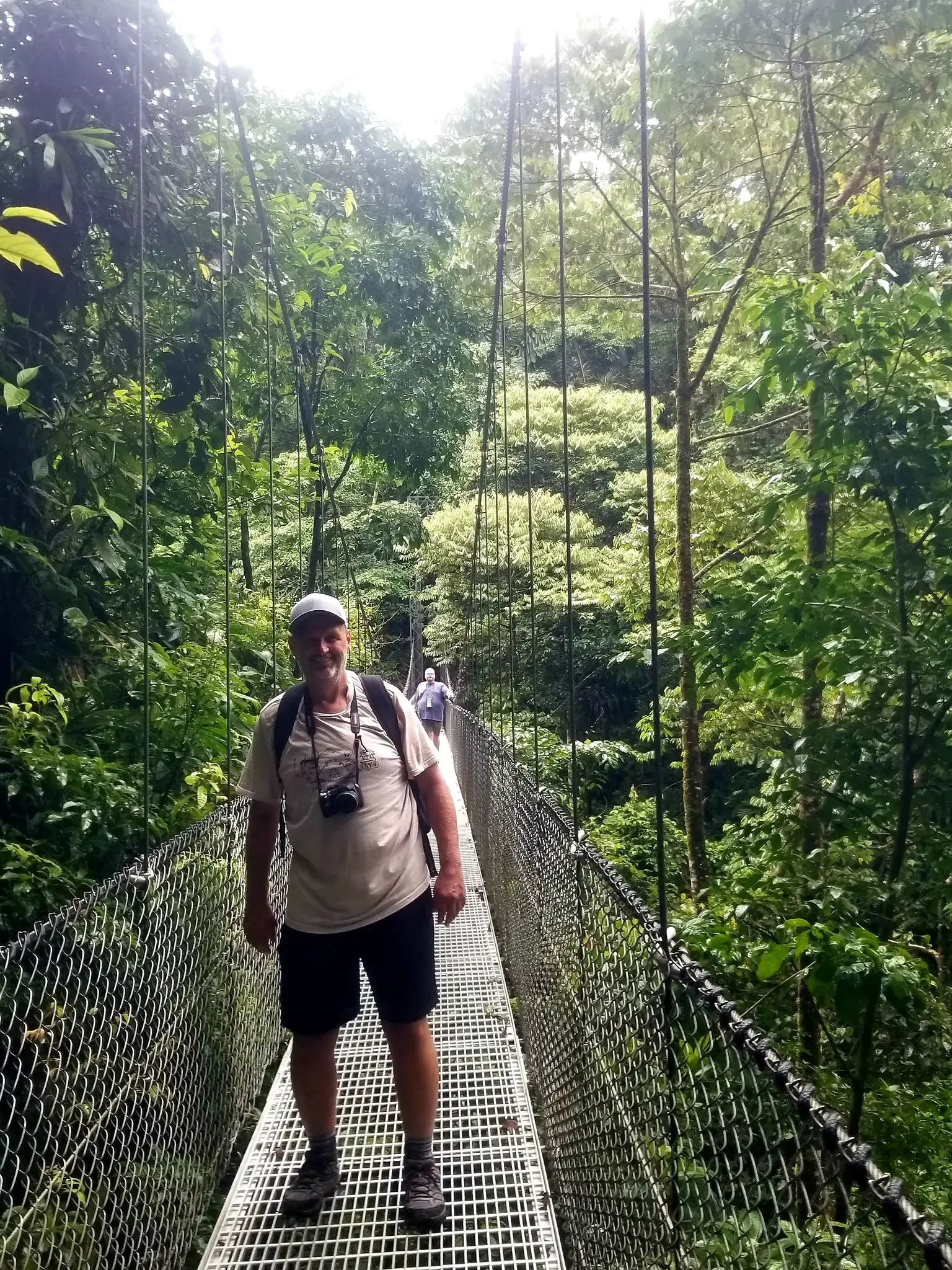 Man standing on a suspension canopy bridge in a lush rainforest in Costa Rica as other hikers cross the bridge.
