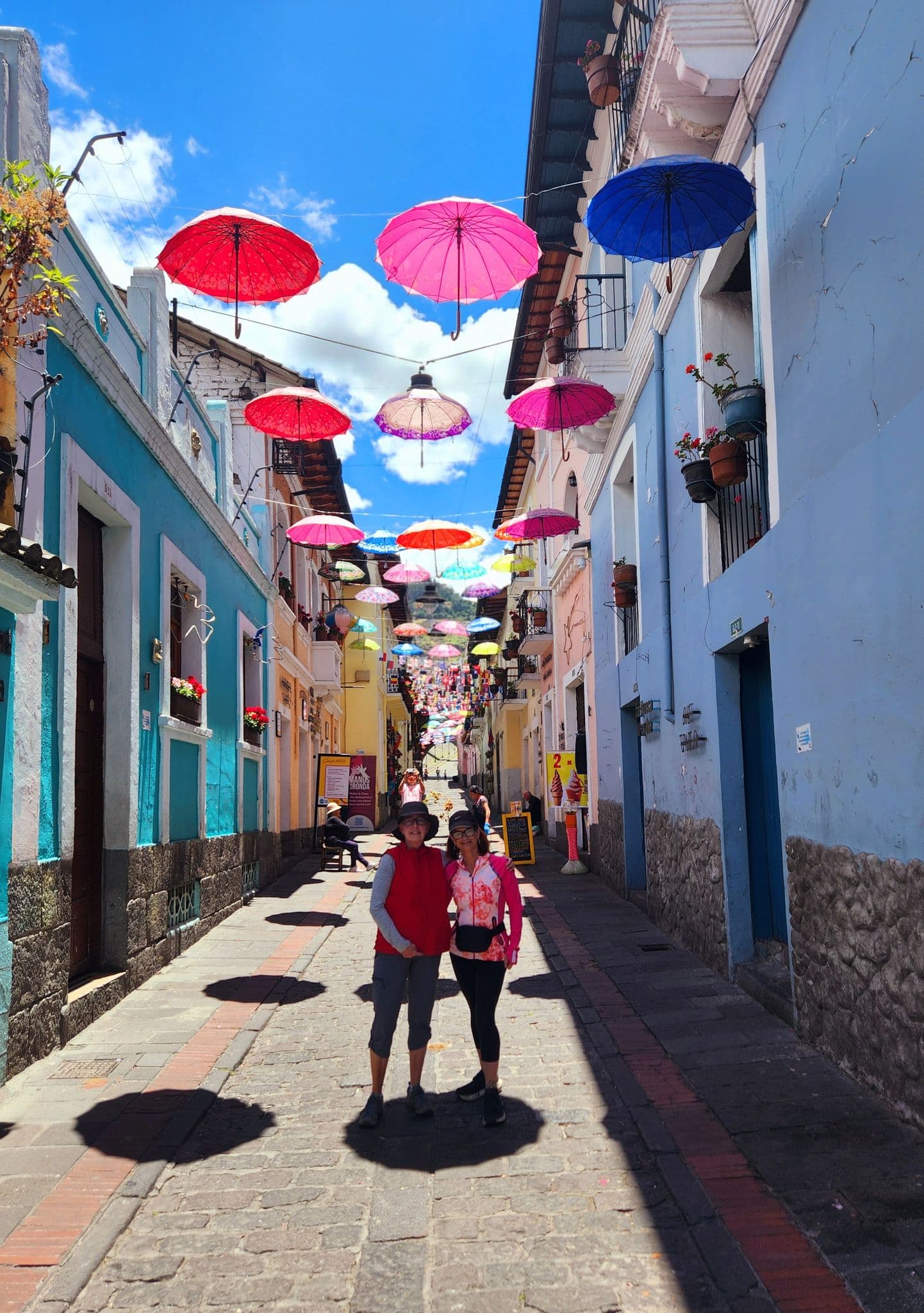 Colorful umbrellas suspended above a narrow street in Quito, Ecuador, with two travelers standing on the cobblestones below.