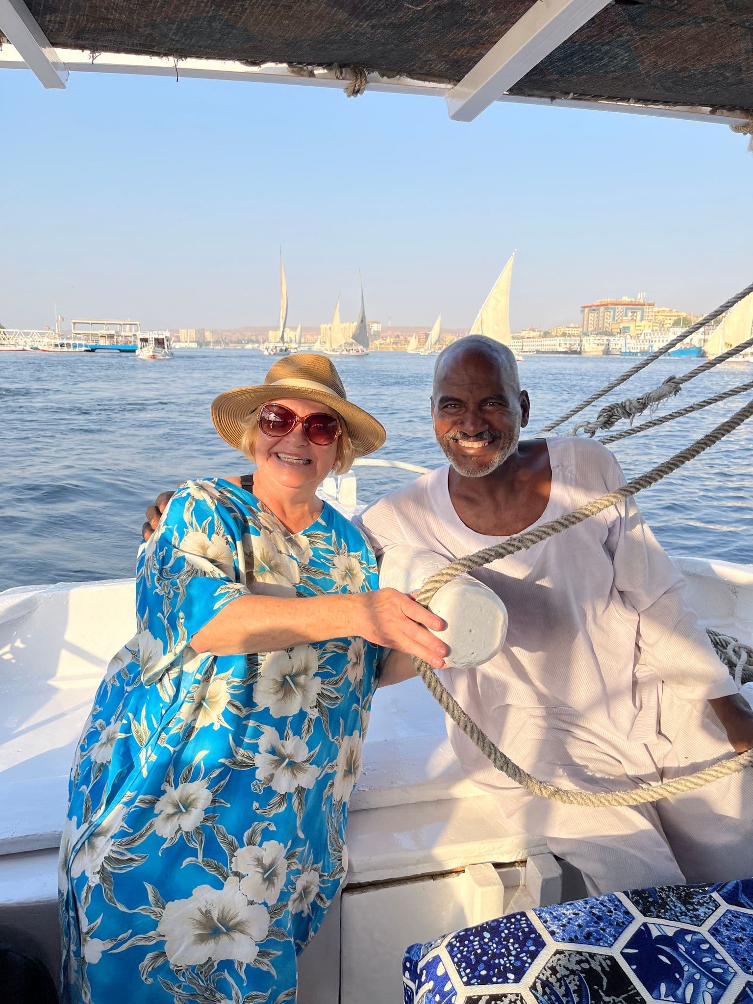 Felucca sailboat on the Nile River with two travelers smiling as one steers the boat, other sails and shoreline visible, Egypt