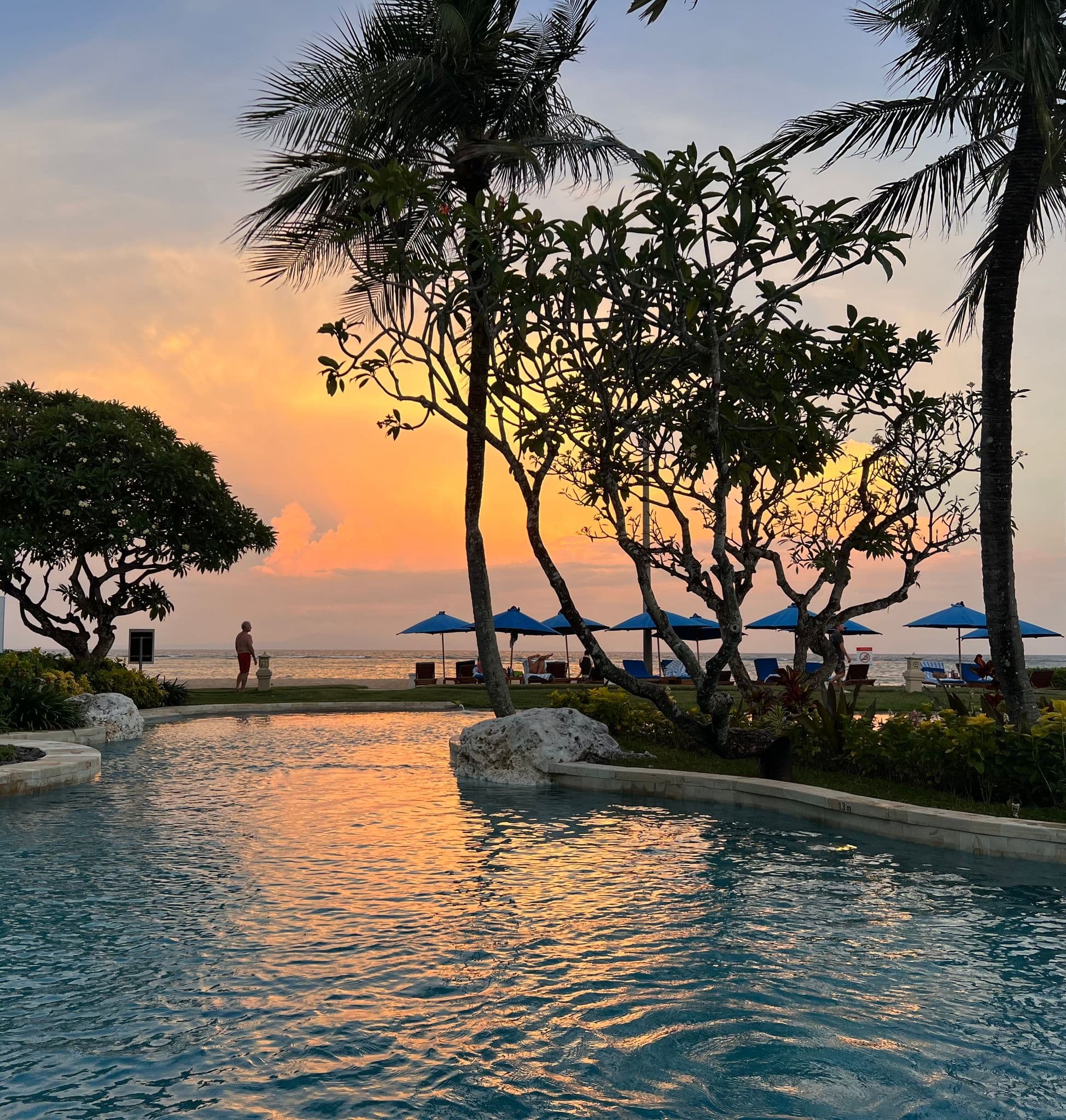 Sunset sky reflecting on a resort pool with palm trees and blue umbrellas at Benoa Beach, Bali, Indonesia.