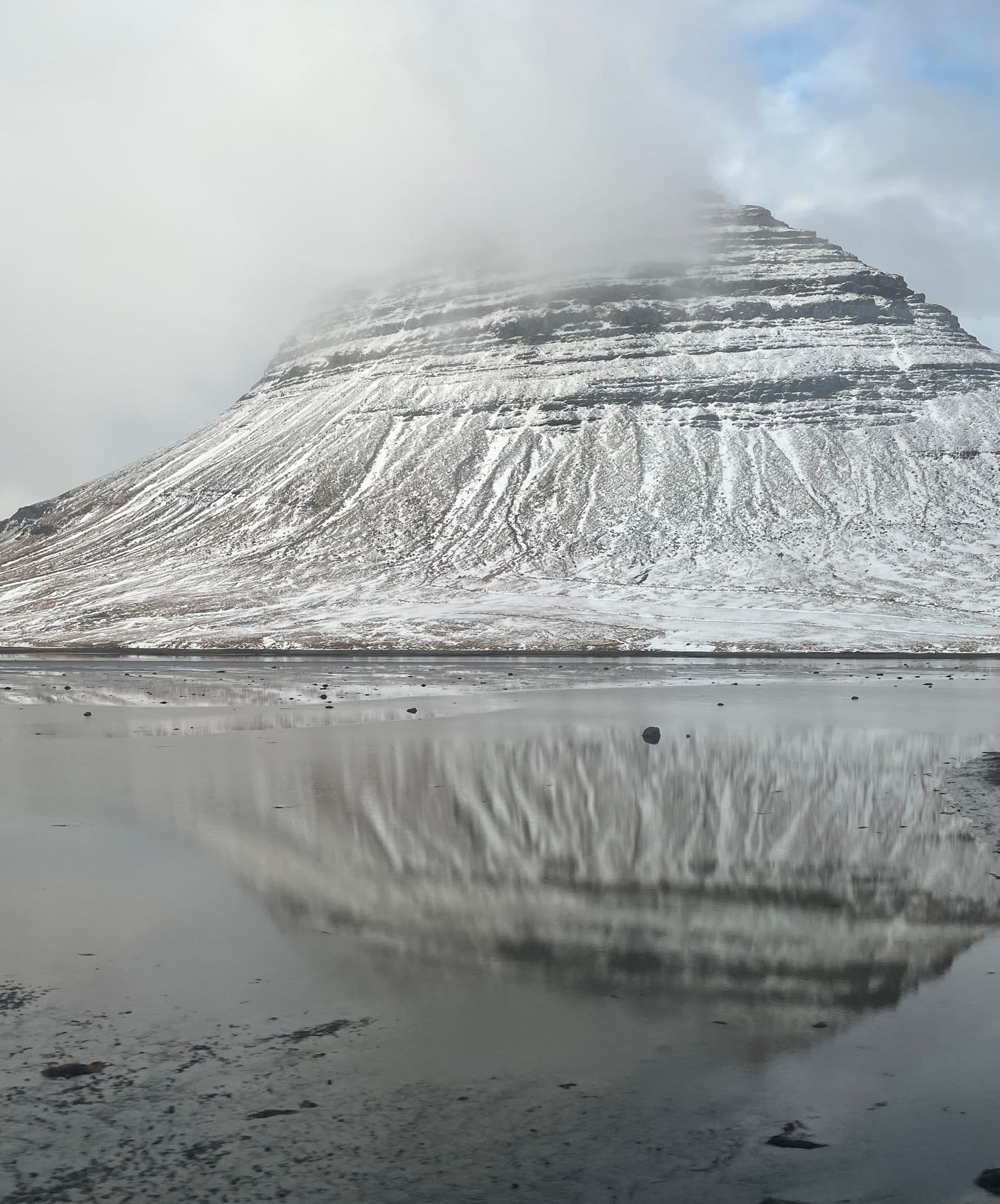 Kirkjufell mountain reflected in calm fjord water, snow-dusted slopes and mist near Grundarfjörður, Iceland.