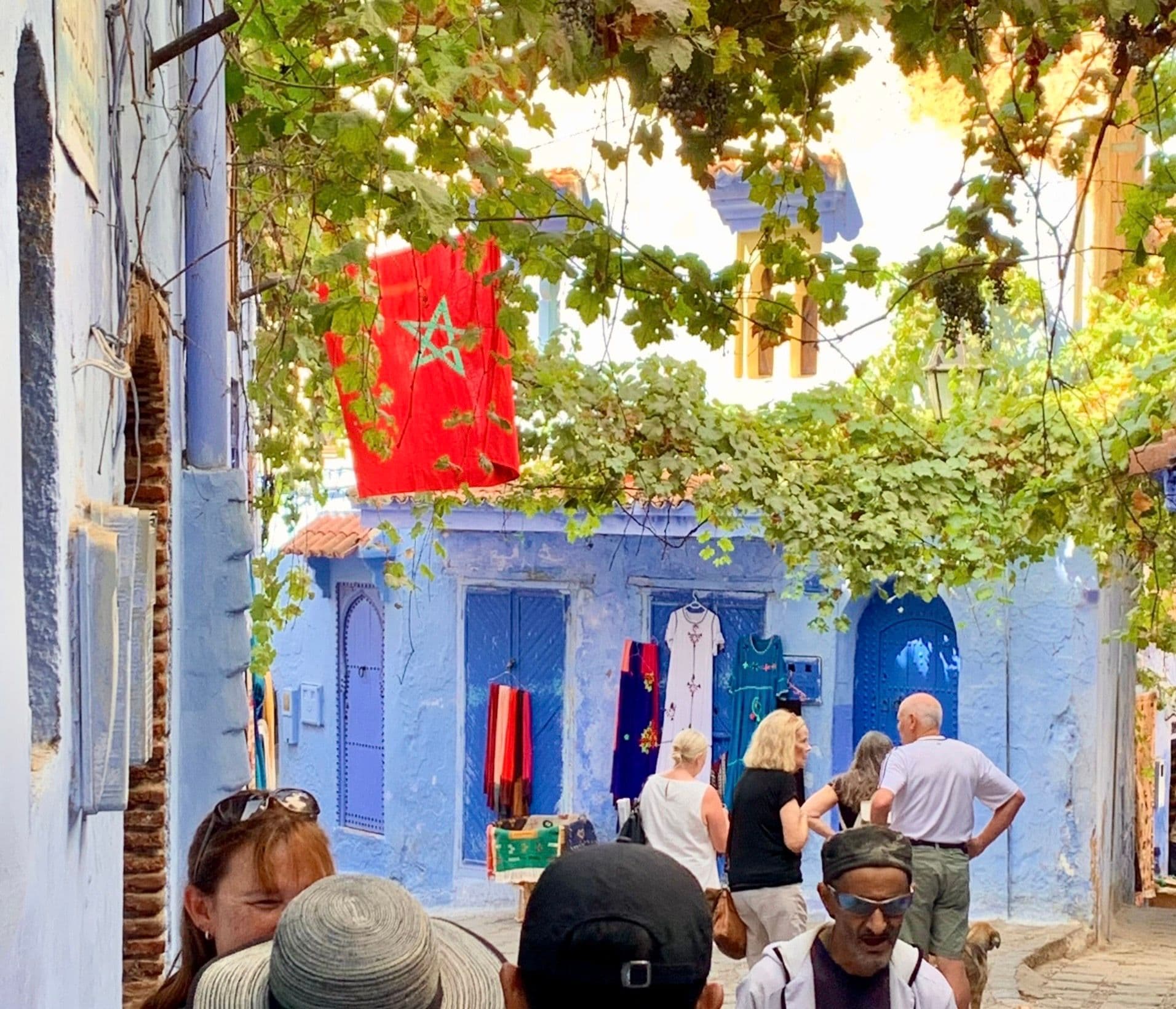 Moroccan flag over blue-washed buildings in the medina of Chefchaouen, Morocco, with tourists walking beneath grapevines.