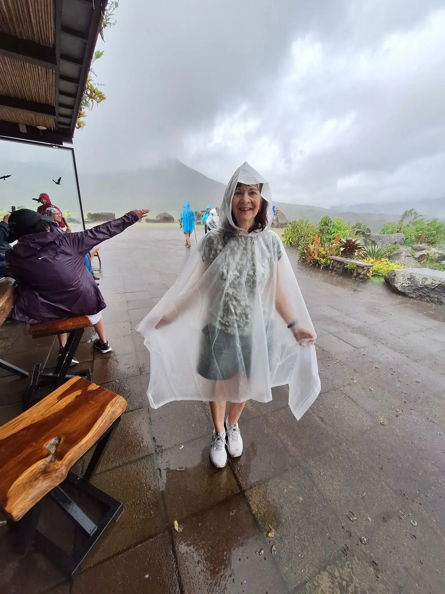 Arenal Volcano partly shrouded in clouds behind a woman in a clear rain poncho standing on a wet viewing area, La Fortuna, Costa Rica.