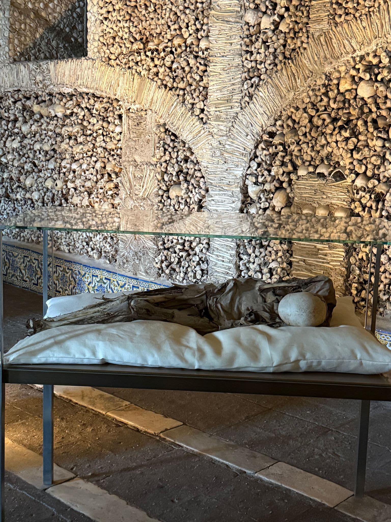 Chapel of Bones interior in Évora, Portugal, walls lined with human skulls and bones and a wrapped corpse displayed under glass