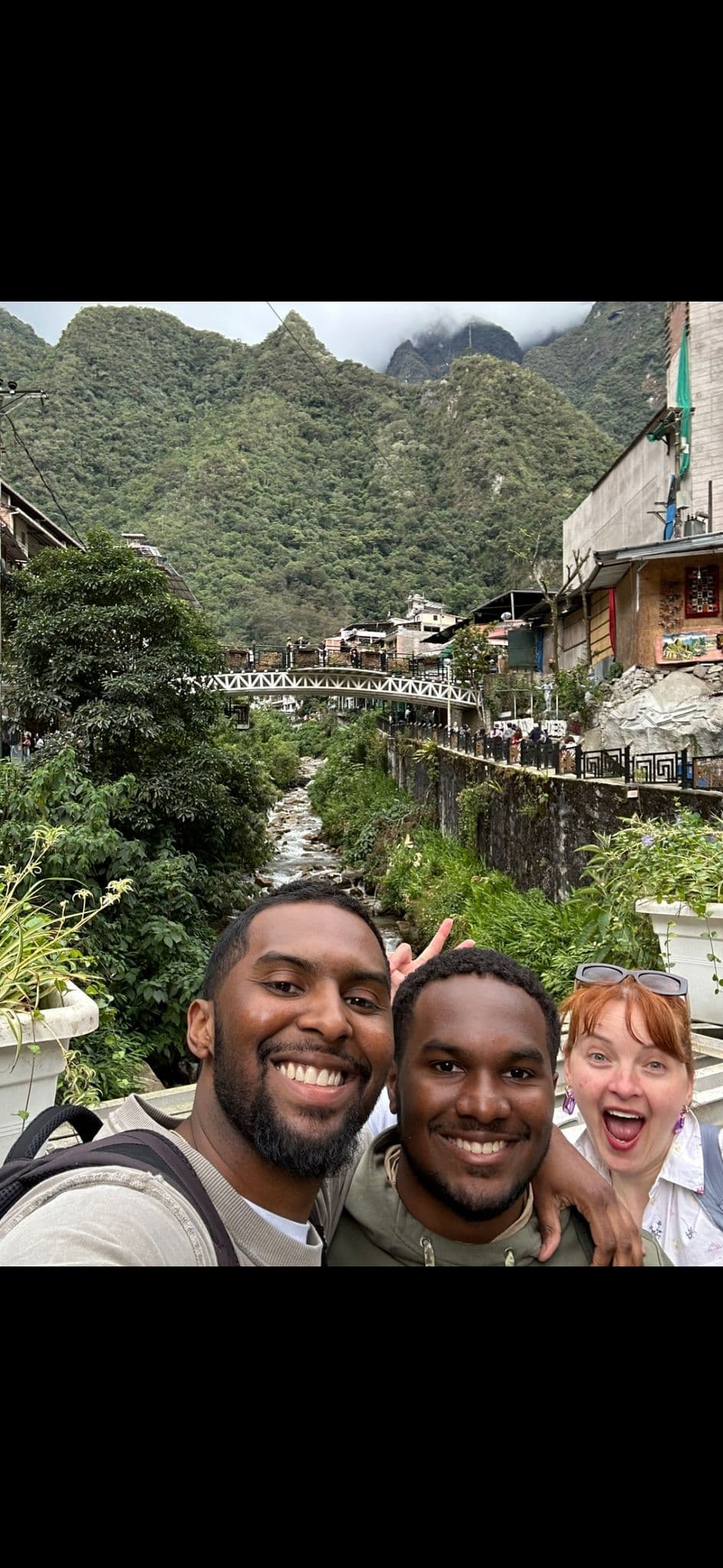 Machu Picchu hillside and river with three travelers taking a smiling selfie in Aguas Calientes, Peru.