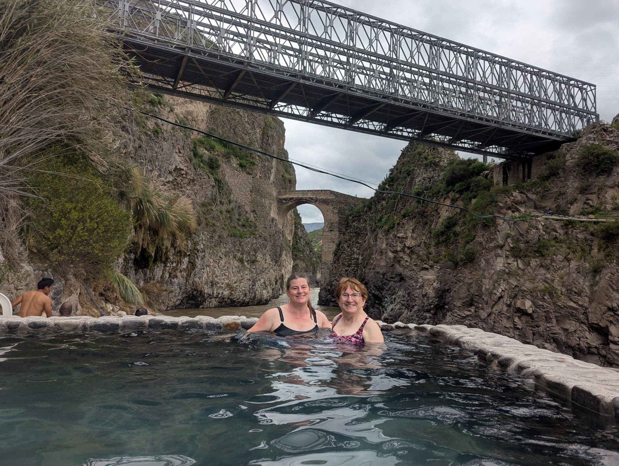 La Calera hot springs pool with two people bathing beneath a metal bridge and stone arch in Colca Canyon, Peru.