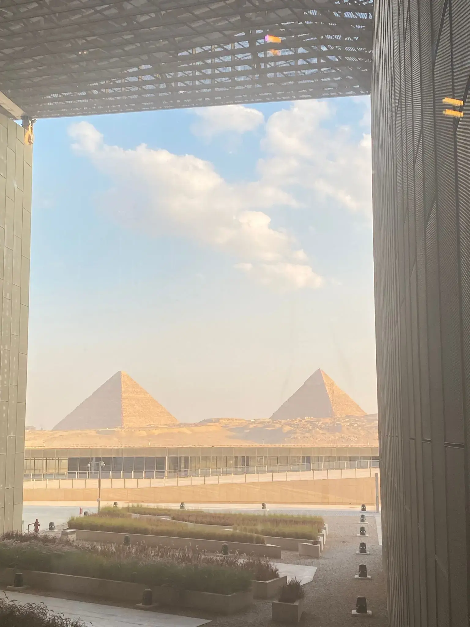 Pyramids of Giza framed through a large Grand Egyptian Museum window with the museum courtyard visible in Giza, Egypt.