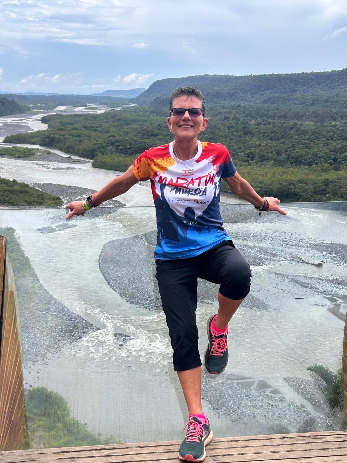Traveler balancing on one leg at a glass viewing platform above a braided river and forested valley.
