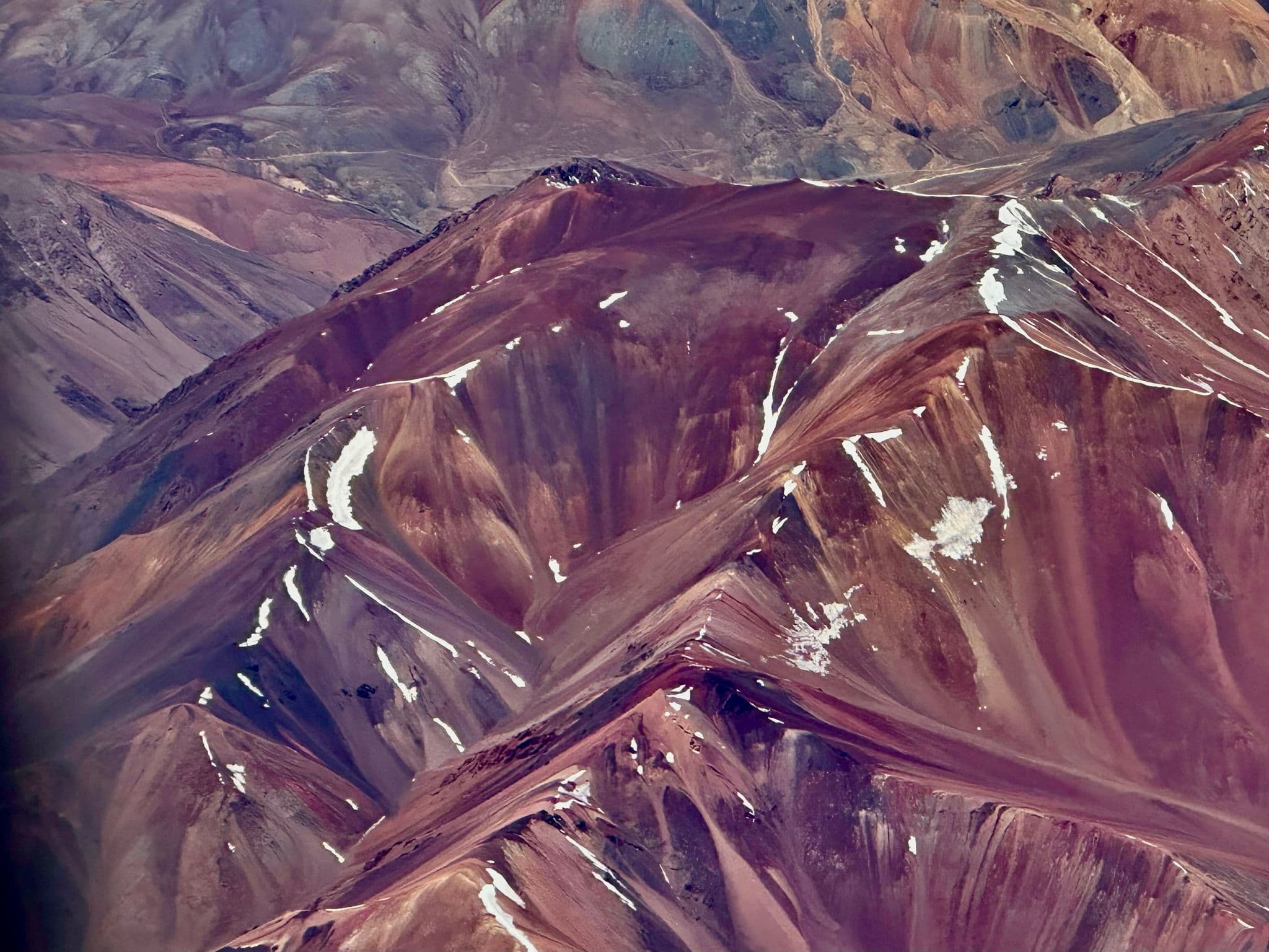 Aerial view of the Andes Mountains with layered reddish peaks and scattered snow patches near the Atacama Desert, Chile.