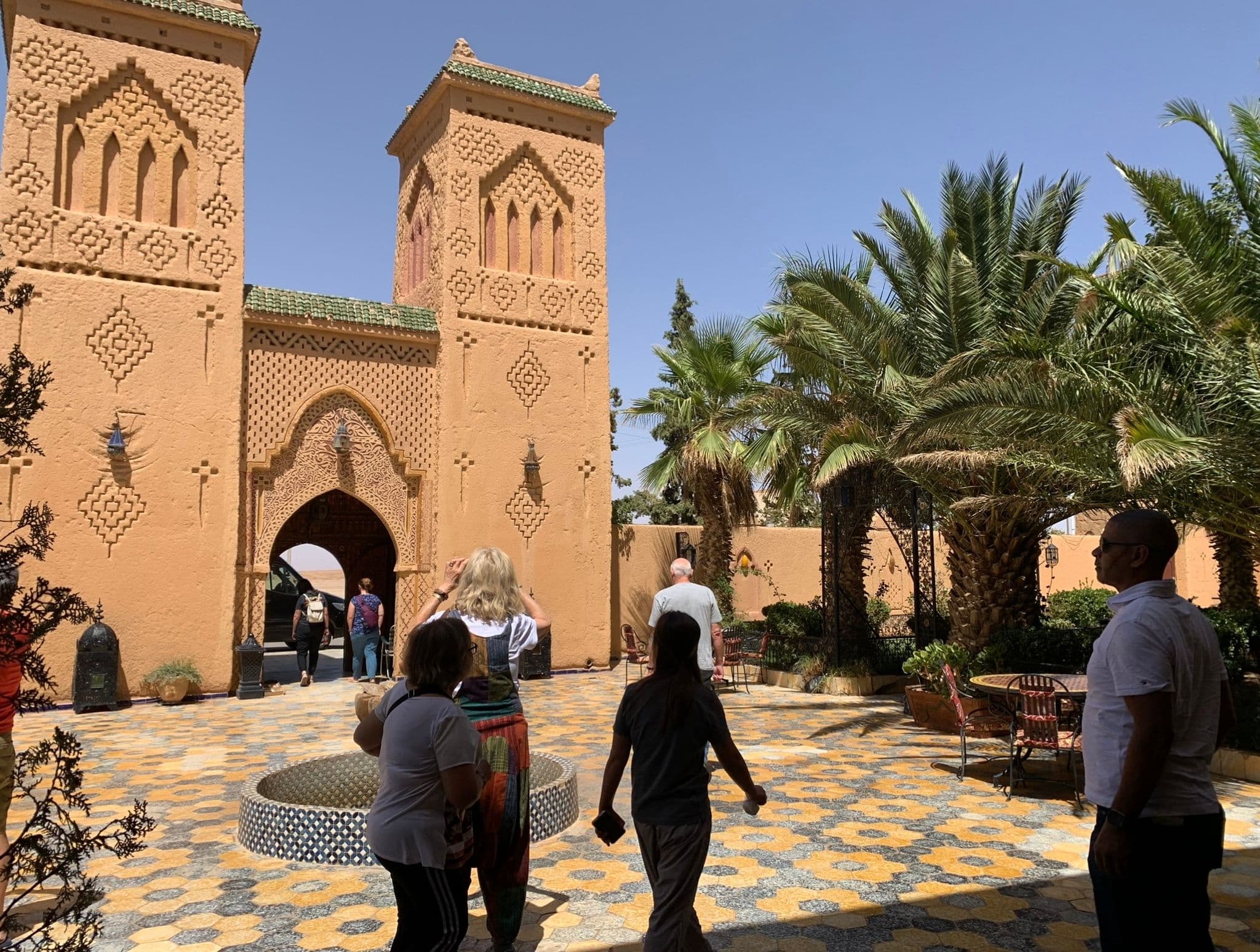 Ornate arched entrance and patterned tile courtyard with palm trees and tourists walking through, Morocco.