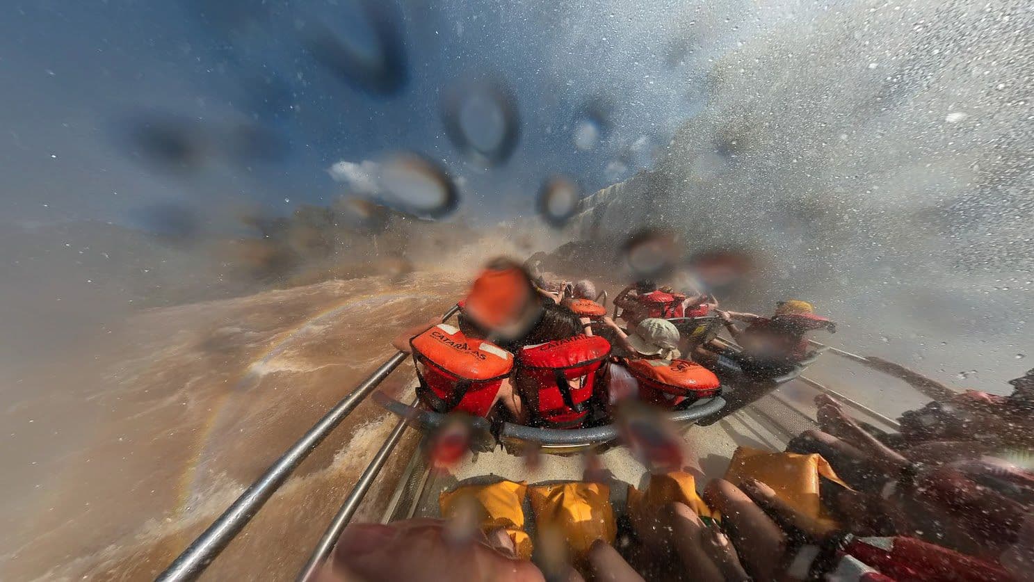 Tour boat with people in orange life jackets approaching Iguazú Falls, Misiones, Argentina, with heavy spray and a faint rainbow.