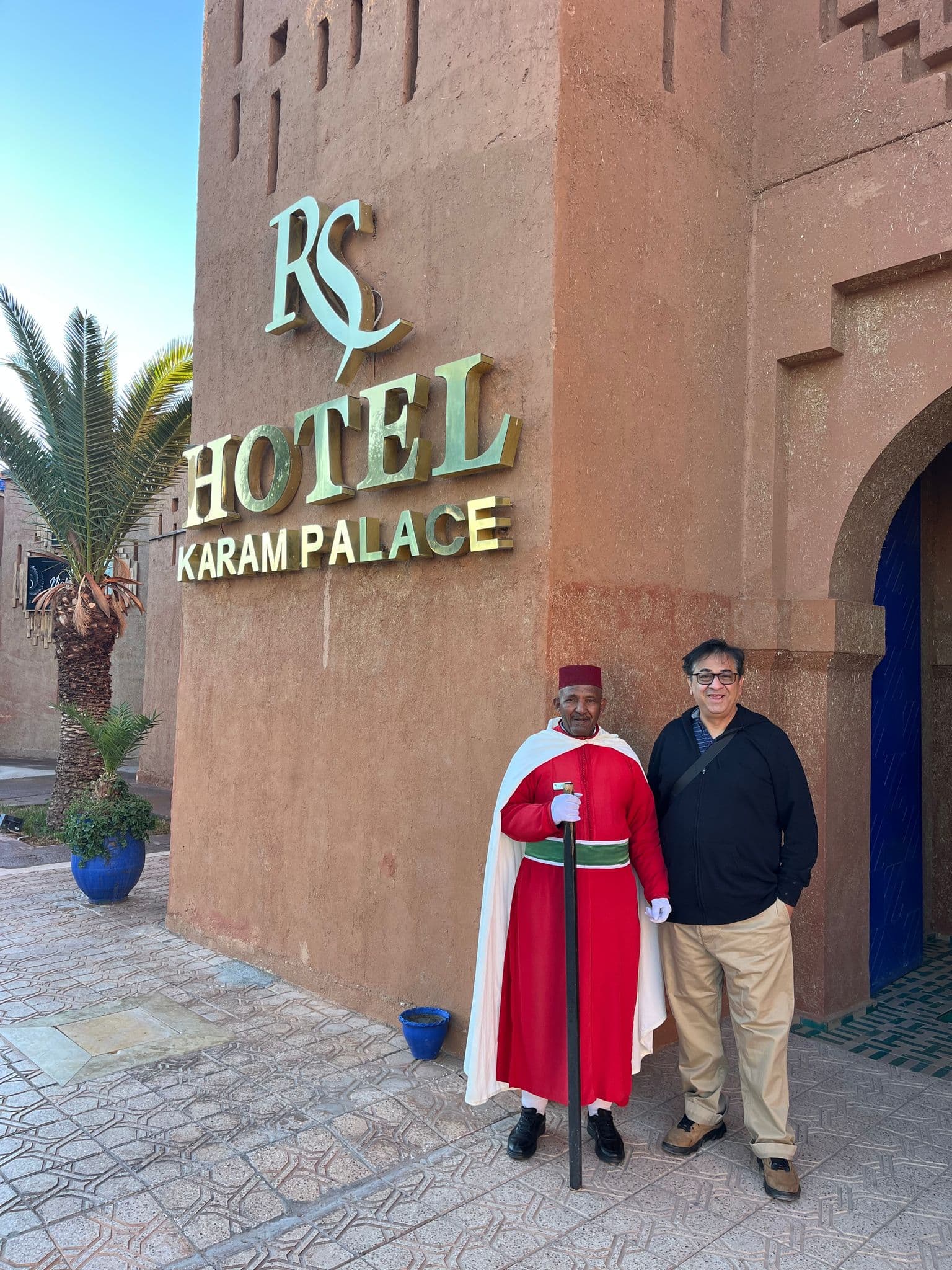 Karam Palace Hotel sign on a terracotta wall with a doorman in a red robe and a traveler posing at the entrance in Morocco.