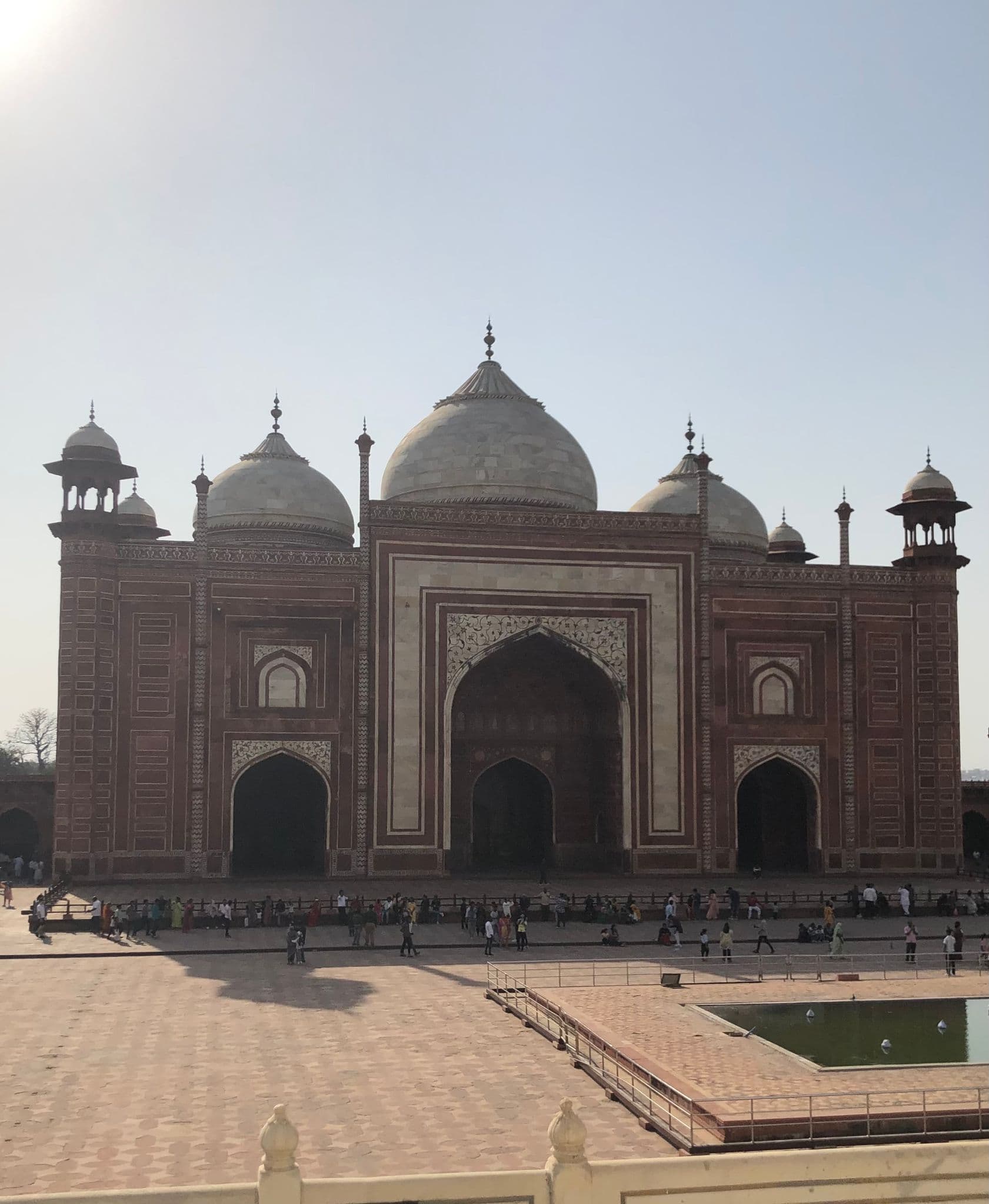 Red sandstone mosque at the Taj Mahal complex in Agra, India, with visitors in the forecourt and a reflecting pool.