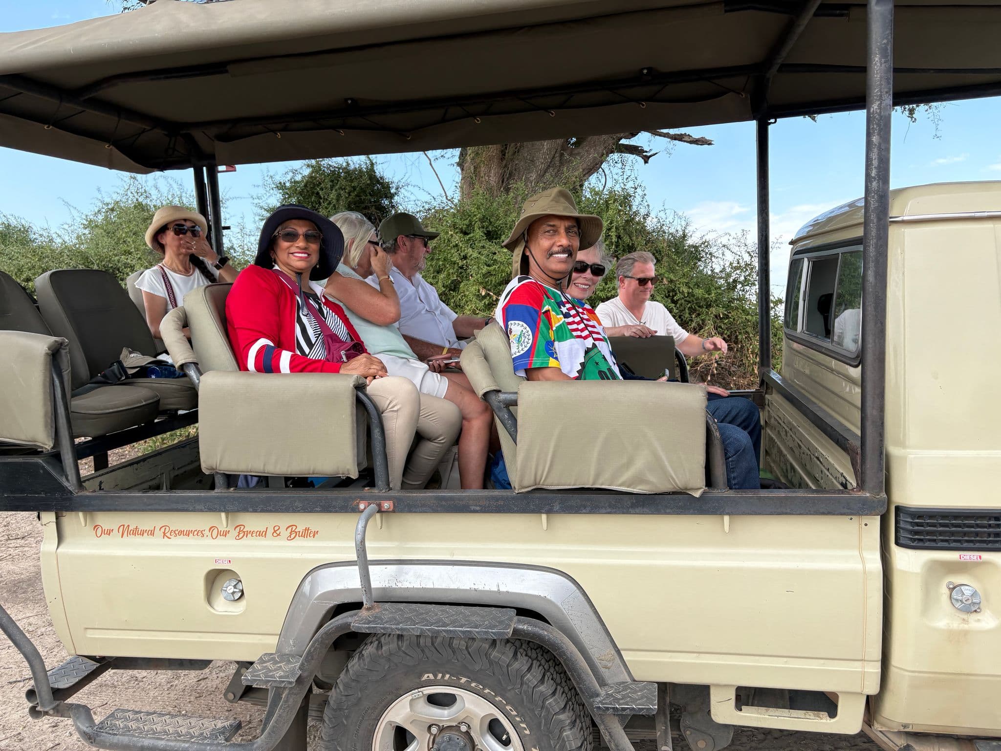 Open safari vehicle with tourists seated and smiling during a game drive in a bush landscape.