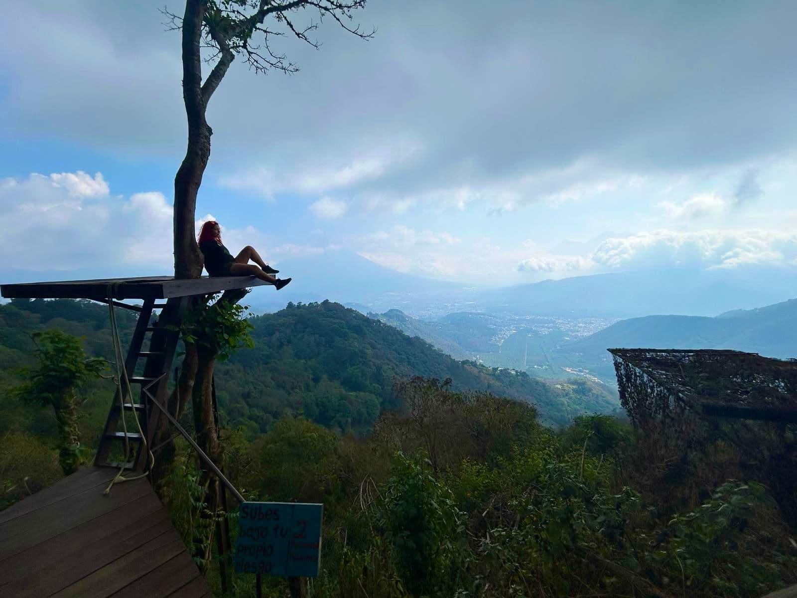 Person sitting on a wooden platform attached to a tree, overlooking a lush valley and a distant volcano in Guatemala.
