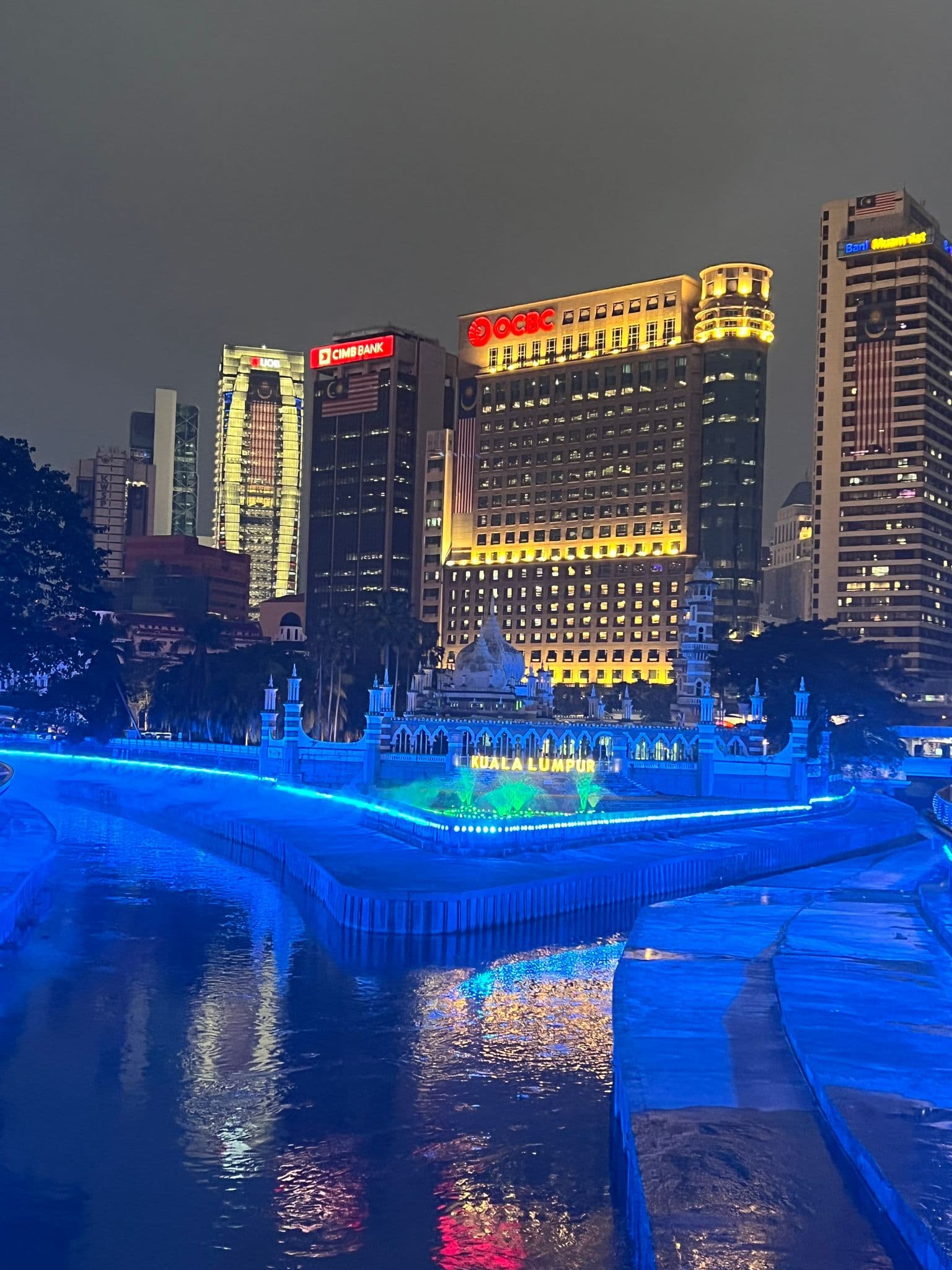 Blue-lit riverside pavilion with KUALA LUMPUR sign and illuminated skyscrapers reflected in a river, Kuala Lumpur, Malaysia