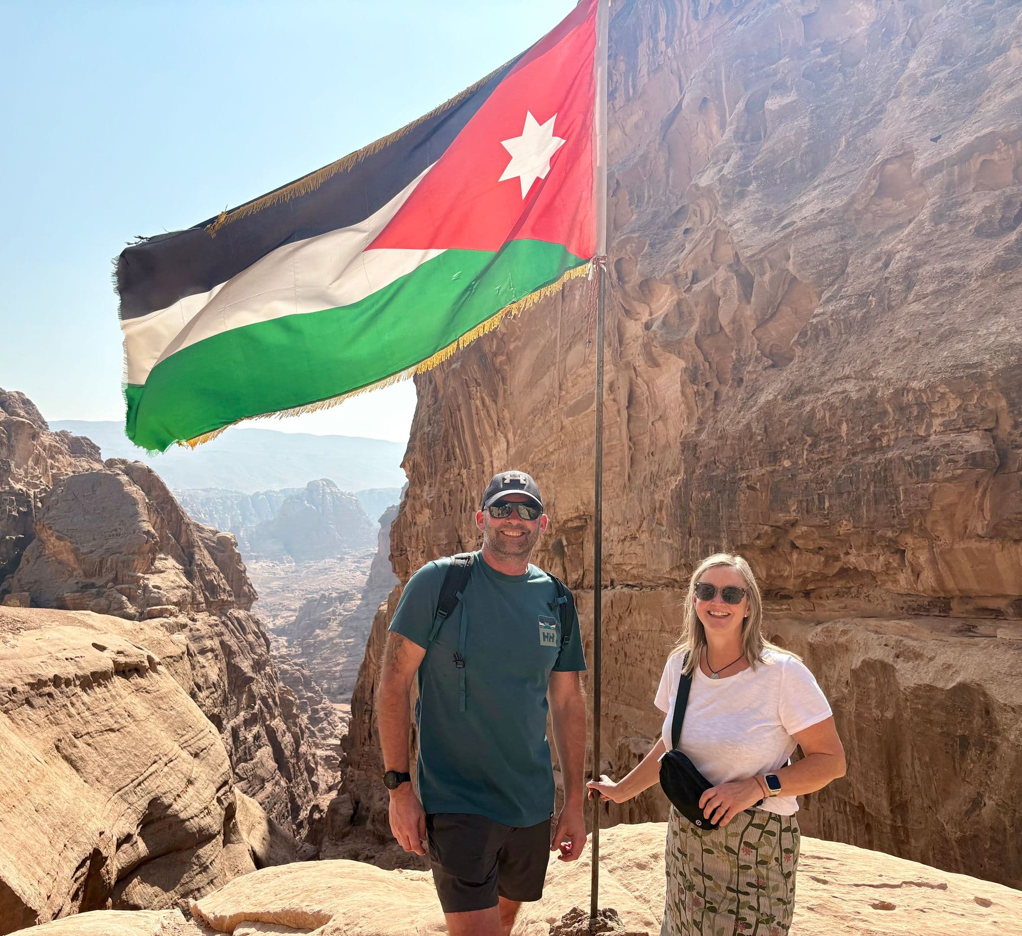 Petra's red sandstone cliffs and a large Jordanian flag with two travelers standing beside the flagpole in Petra, Jordan.