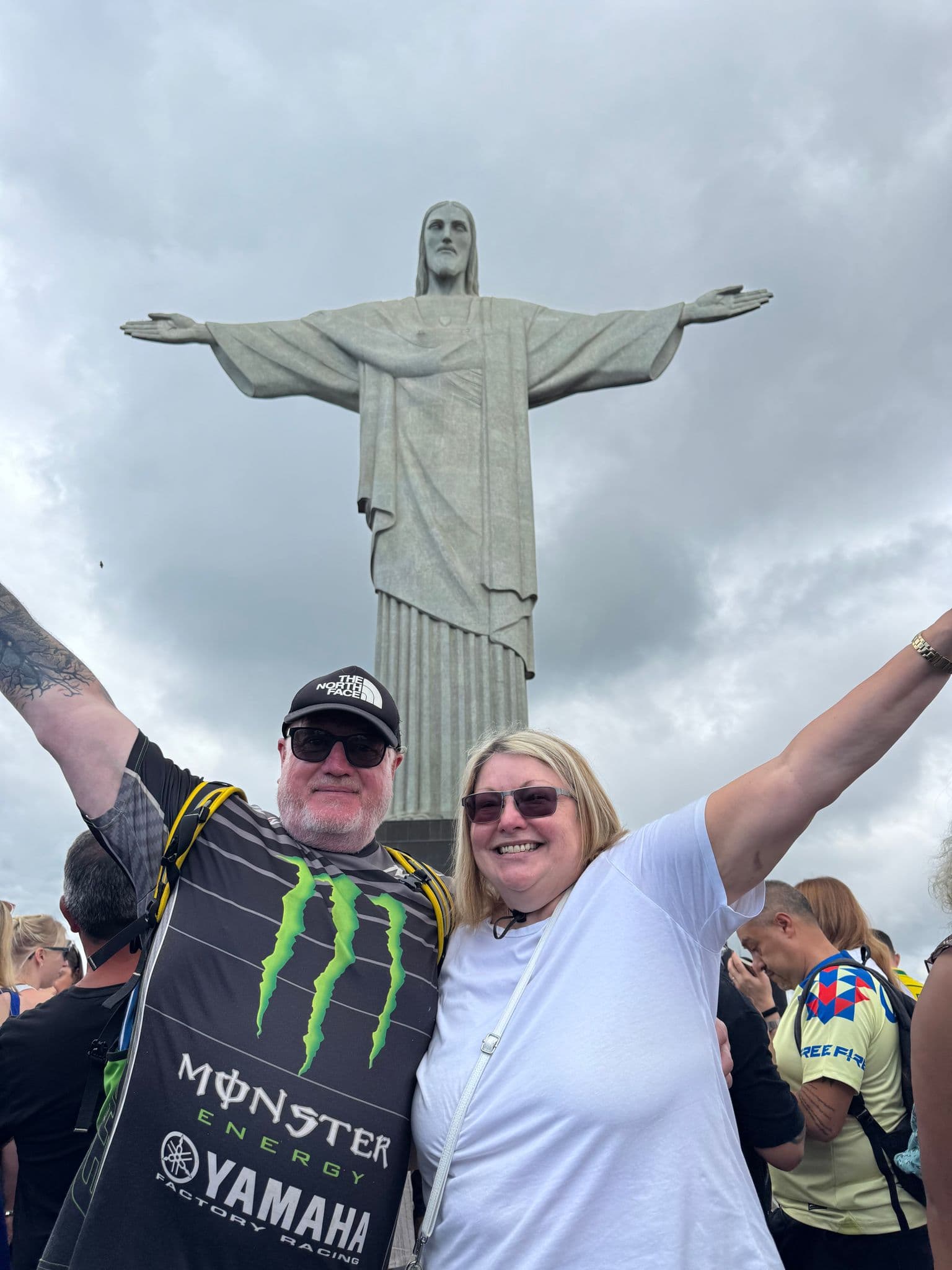 Christ the Redeemer statue with two visitors raising their arms at a crowded viewpoint, Rio de Janeiro, Brazil.