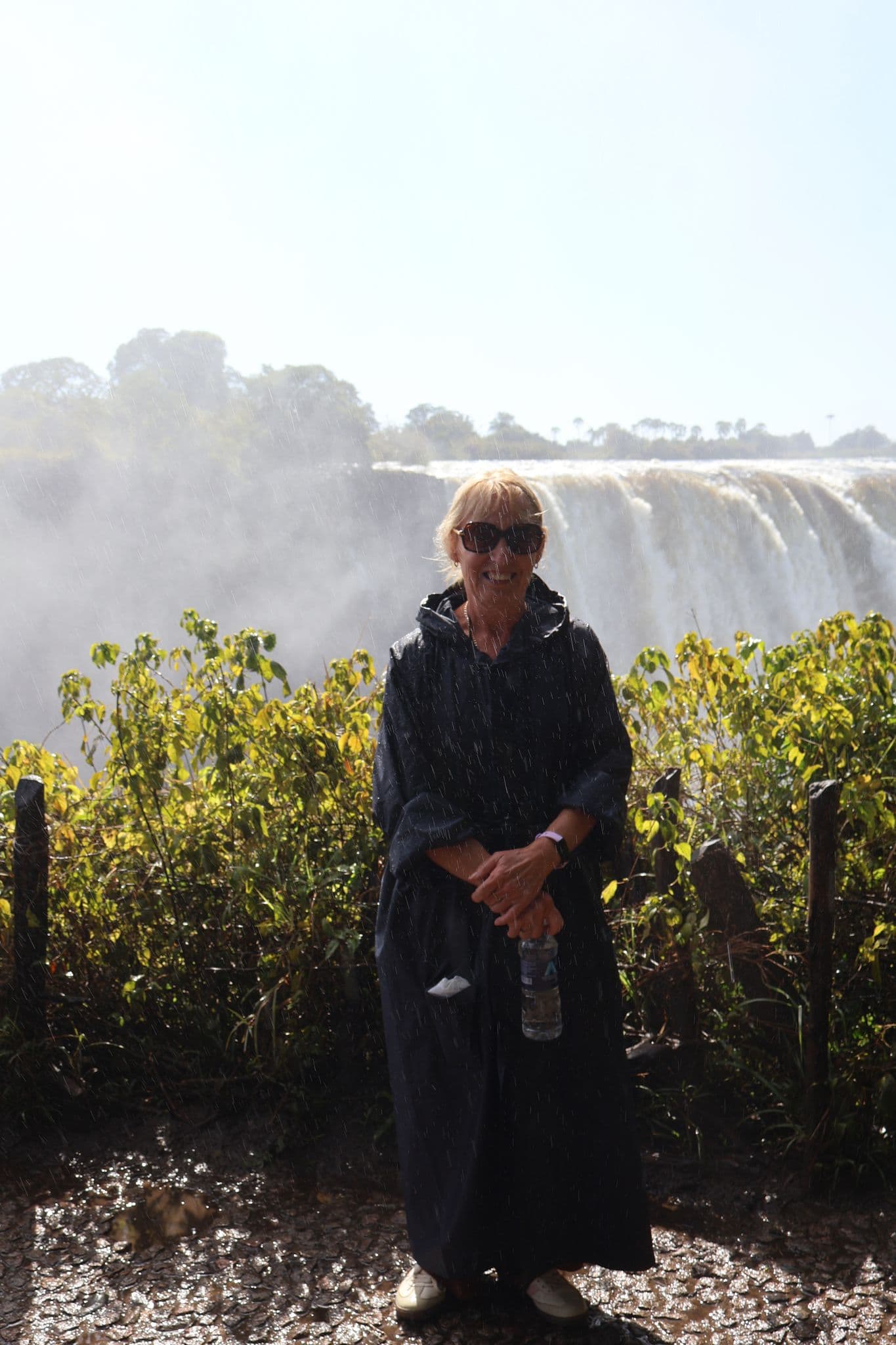 Victoria Falls in the background with a traveler in a dark rain poncho standing near the mist and holding a water bottle.