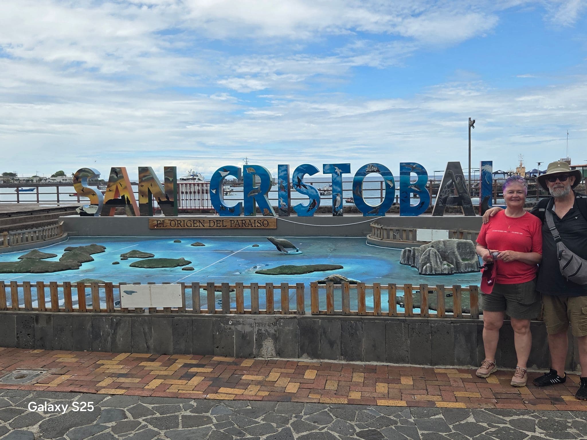 San Cristóbal sign with two travelers posing beside it on the waterfront in Puerto Baquerizo Moreno, Galápagos, Ecuador.