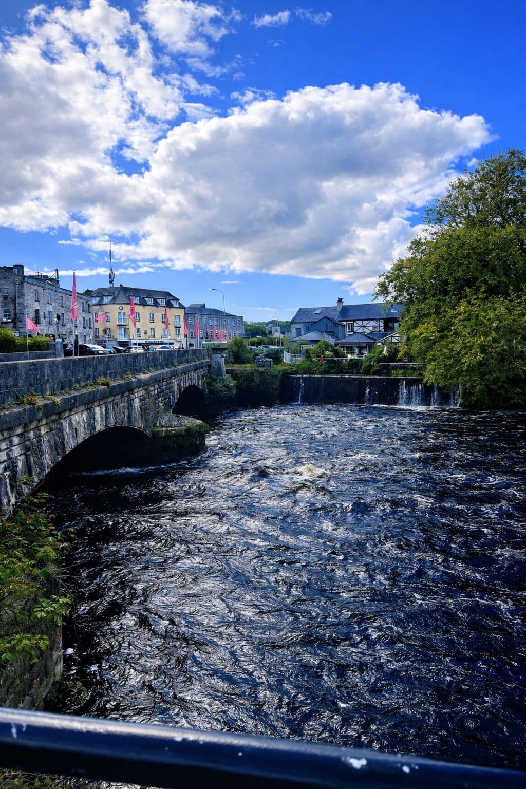 River Corrib flowing under a stone bridge with riverside buildings and a small waterfall, Ireland.