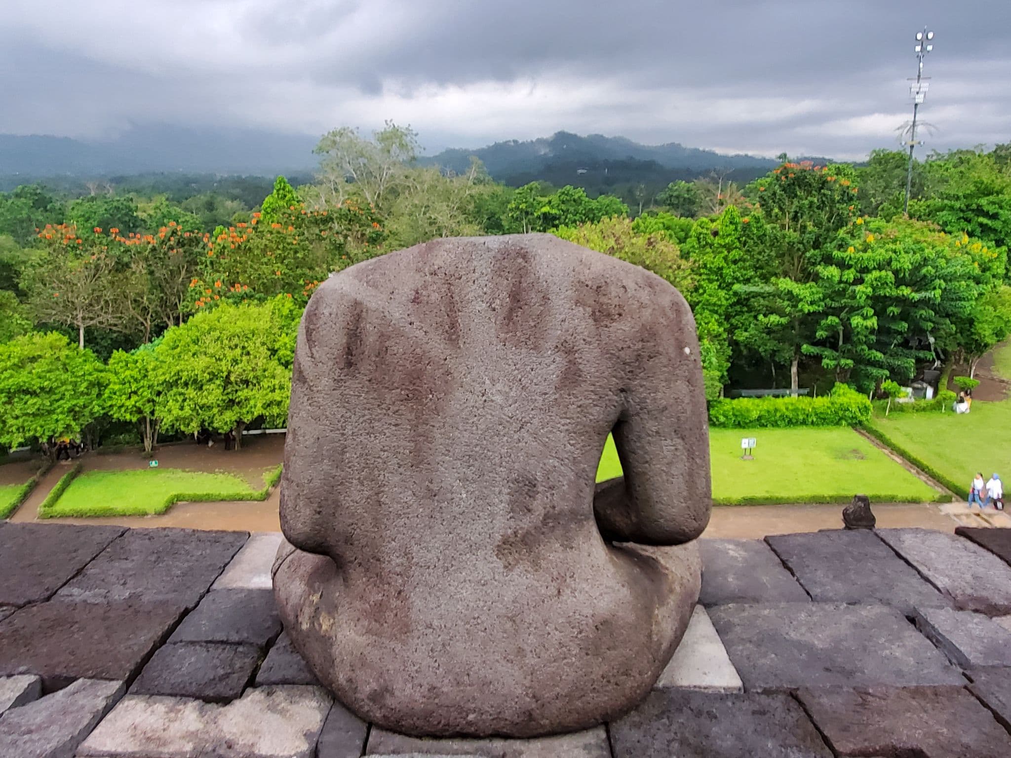 Back of a headless stone Buddha statue at Borobudur temple, overlooking tropical trees and distant hills in Central Java, Indonesia.