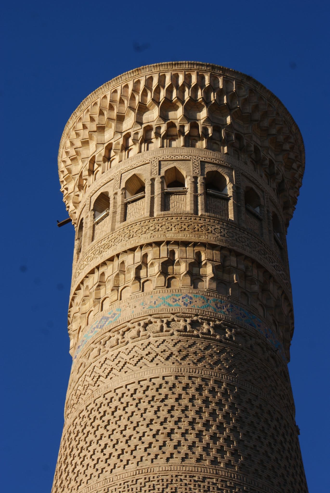 Islom Hoja Minaret in Khiva, Uzbekistan, seen from below against a clear blue sky, showing carved brickwork and a turquoise tile band.