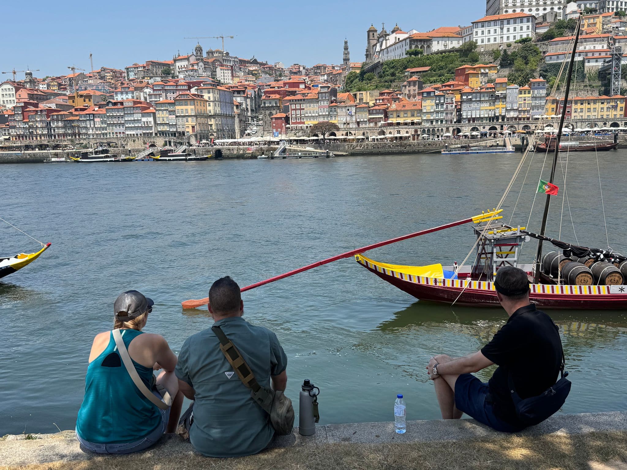 Rabelo boat on the Douro River at the Ribeira waterfront in Porto, Portugal, with three people sitting on the riverbank.